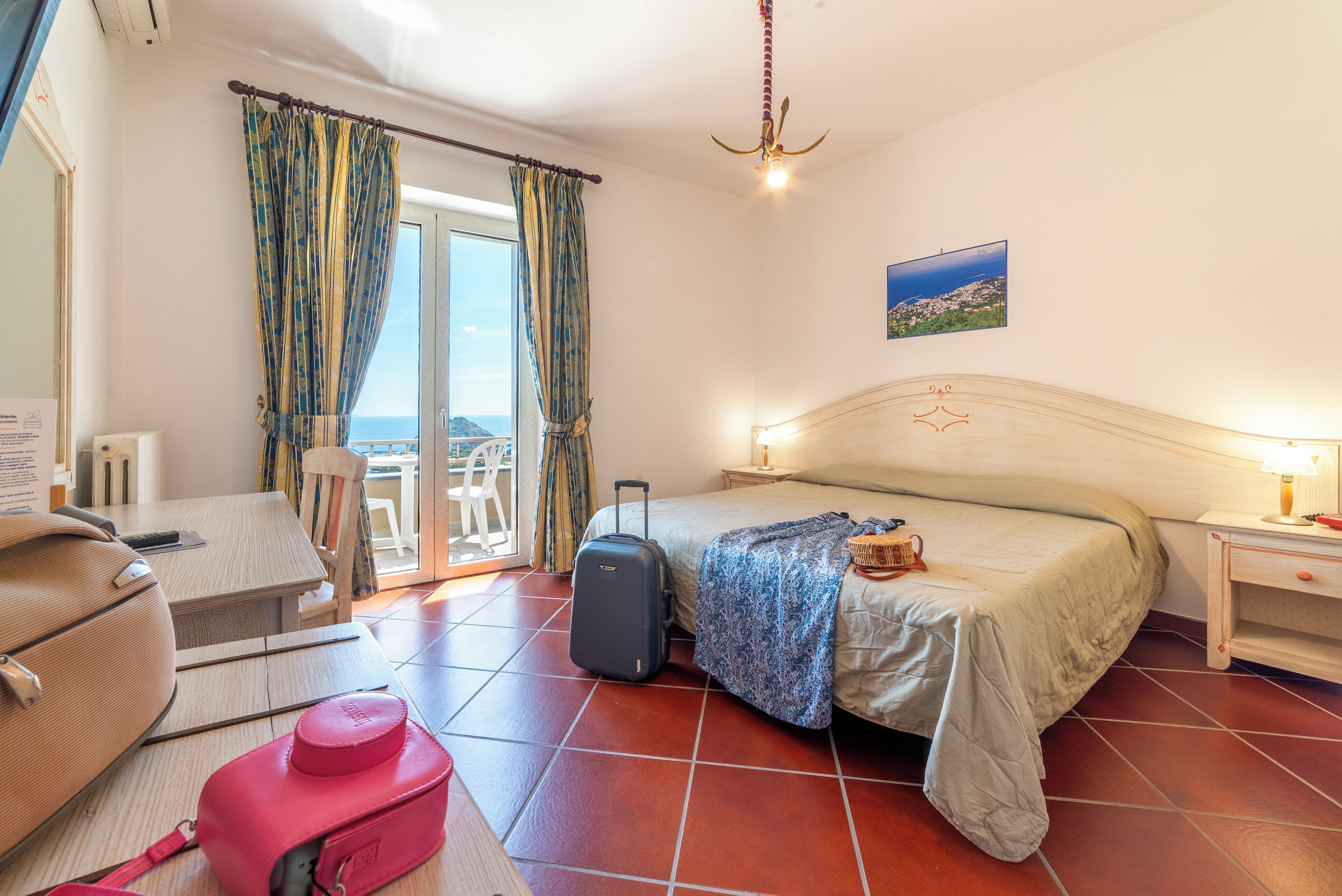 A hotel room with a bed, a chair, a table, a suitcase, and a suitcase on wheels.