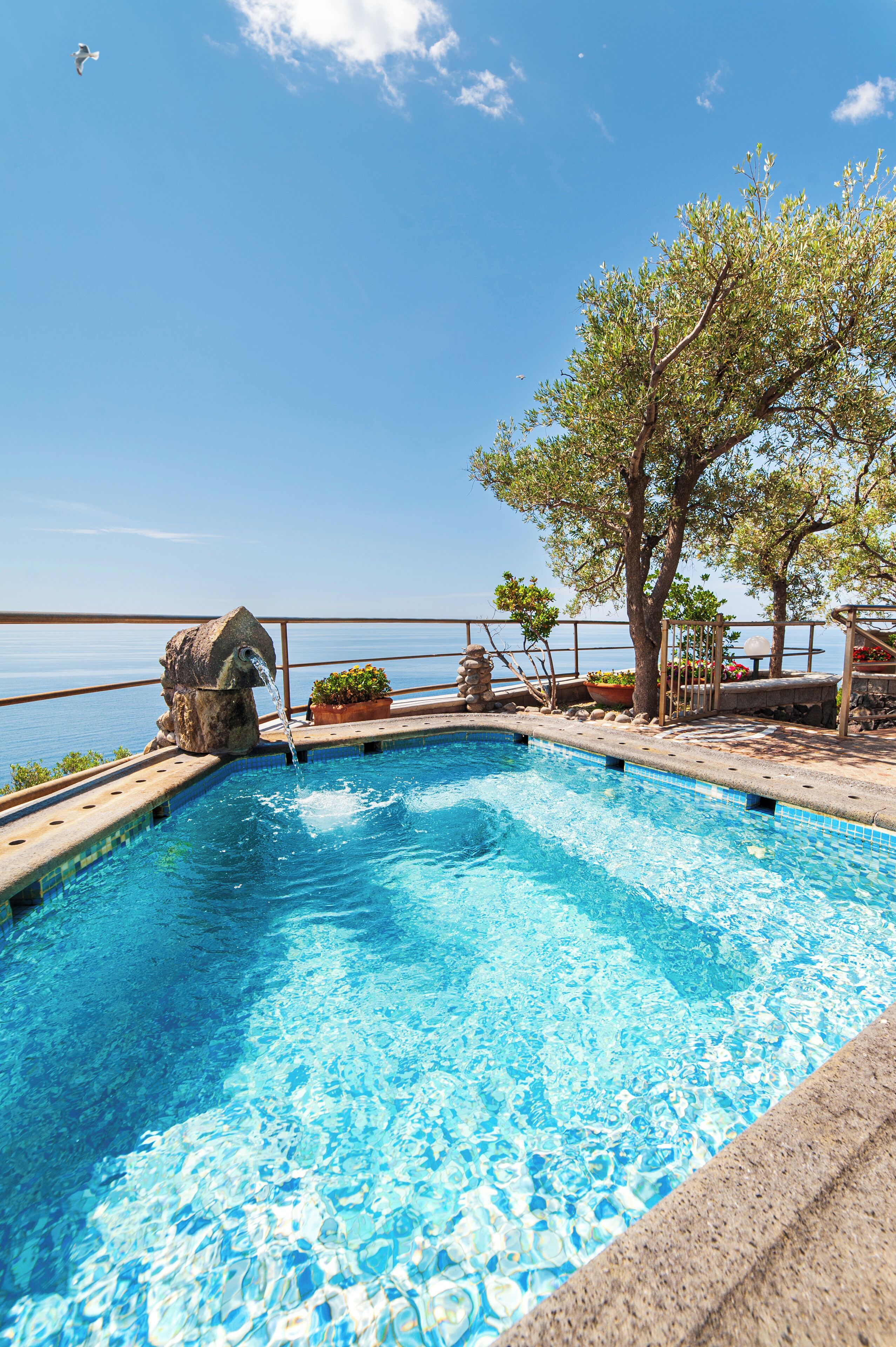 A swimming pool with a stone fountain, surrounded by potted plants and a view of the sea.
