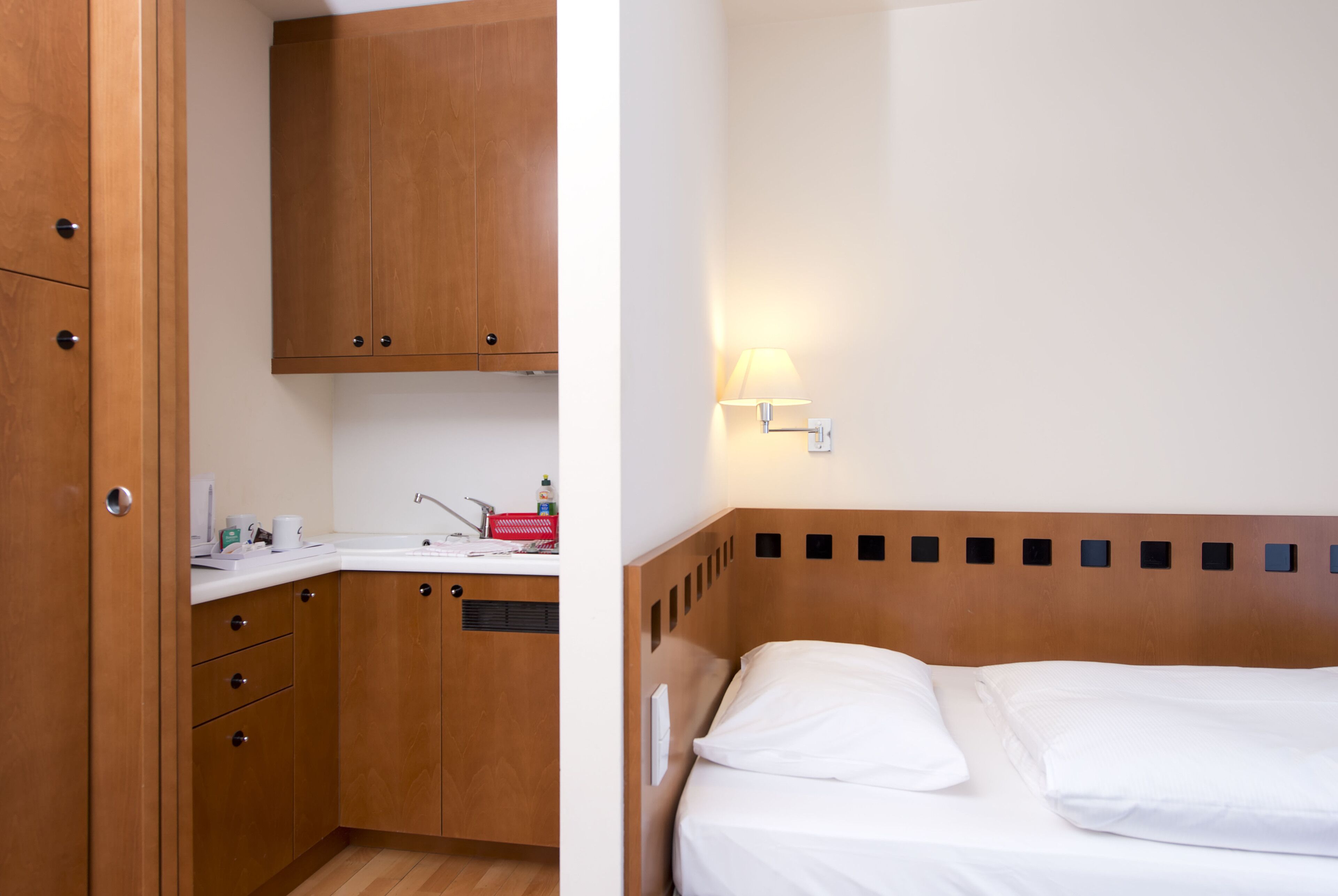 Una habitación de hotel compacta con un armario de madera, una pequeña kitchenette y una cama individual con ropa blanca.