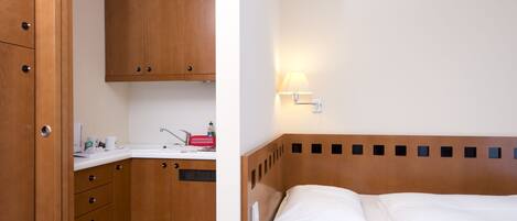 Una habitación de hotel compacta con un armario de madera, una pequeña kitchenette y una cama individual con ropa blanca.