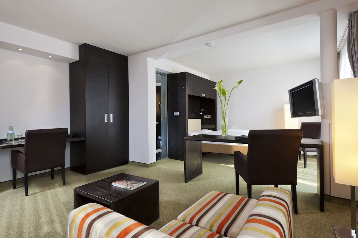 A modern hotel room with a sofa, a coffee table, a desk with a computer, a TV, and a large wardrobe.
