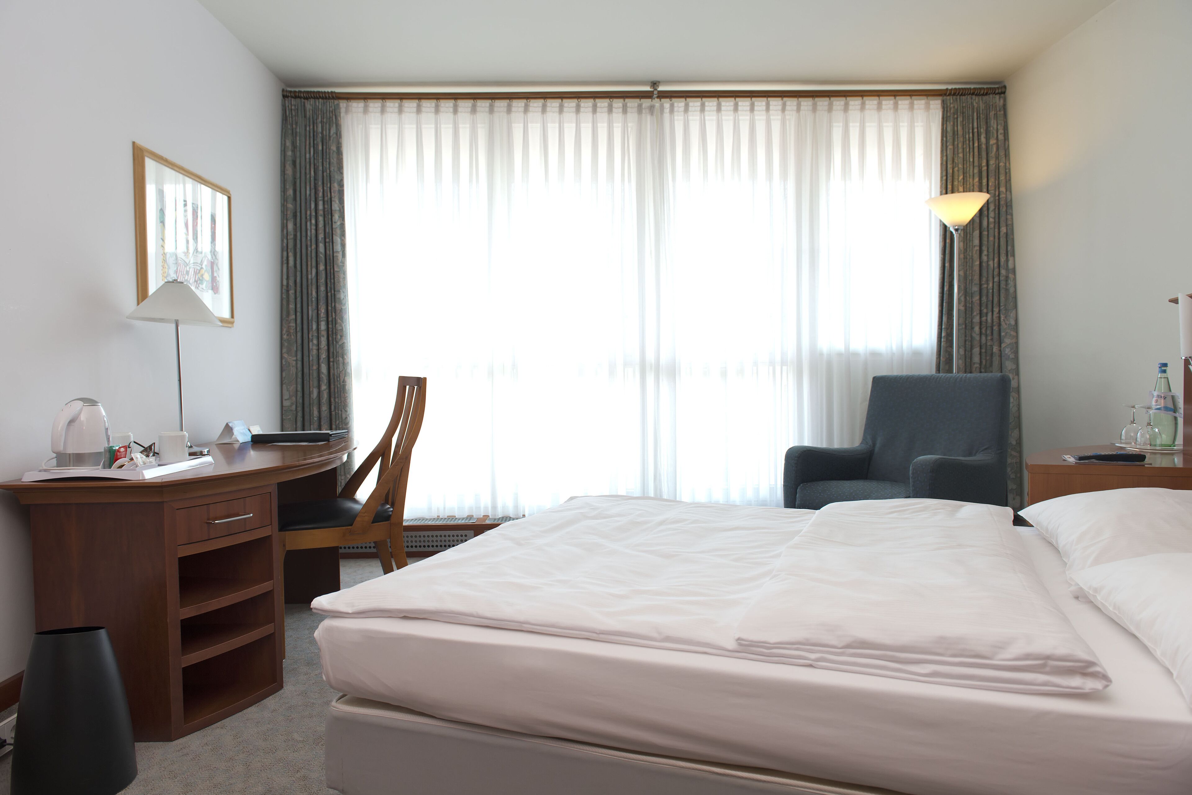 Ein Hotelzimmer mit Bett, Schreibtisch, Stuhl, Lampe und einem Fenster mit Vorhängen.