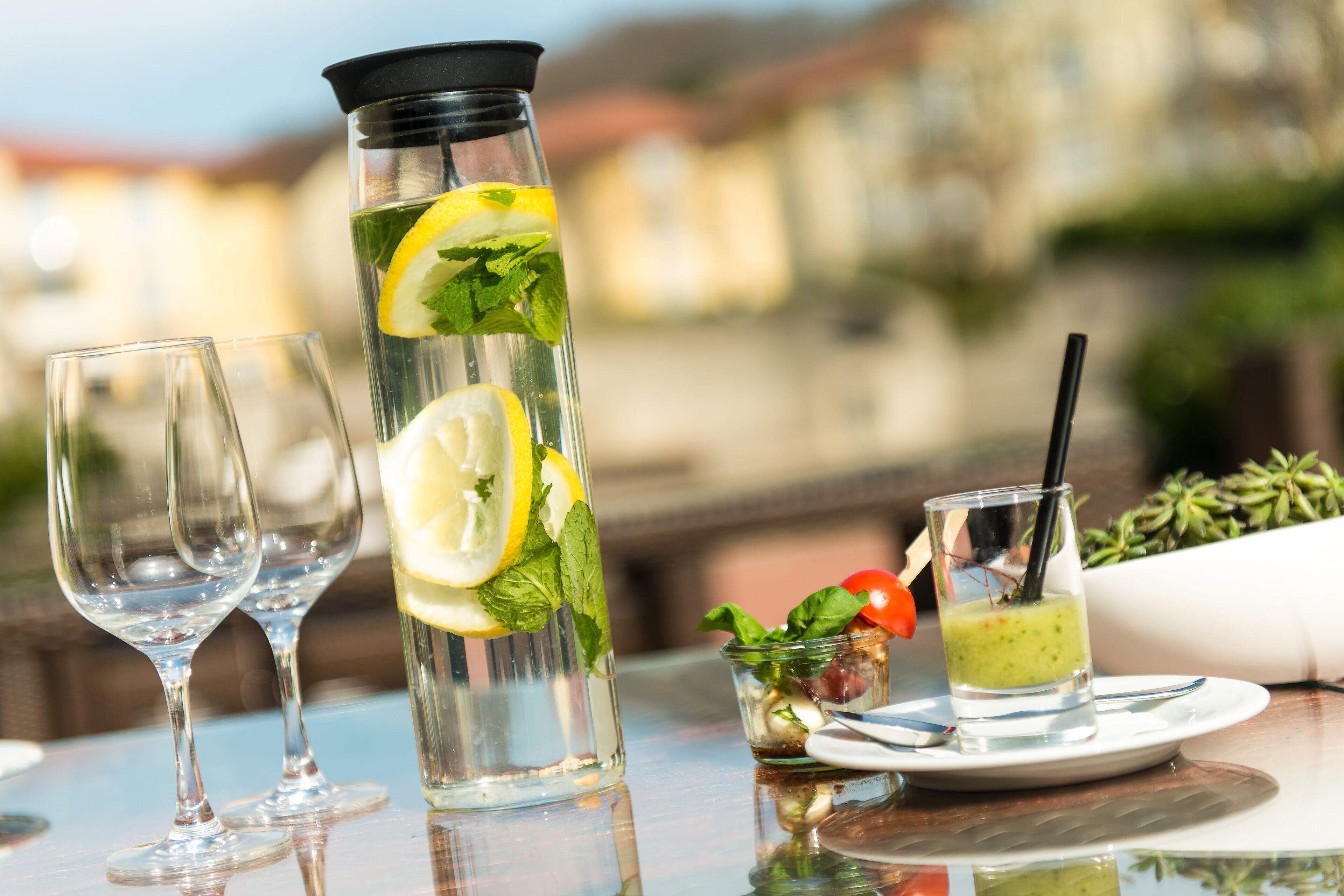 Ein Tisch mit einem hohen Glas, das eine klare Flüssigkeit enthält, dekoriert mit Zitronenscheiben und Minzblättern, sowie einem kleineren Glas mit einem grünen Getränk und einem Strohhalm.