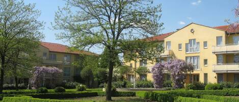 A yellow building with multiple balconies, surrounded by well-maintained hedges and trees.