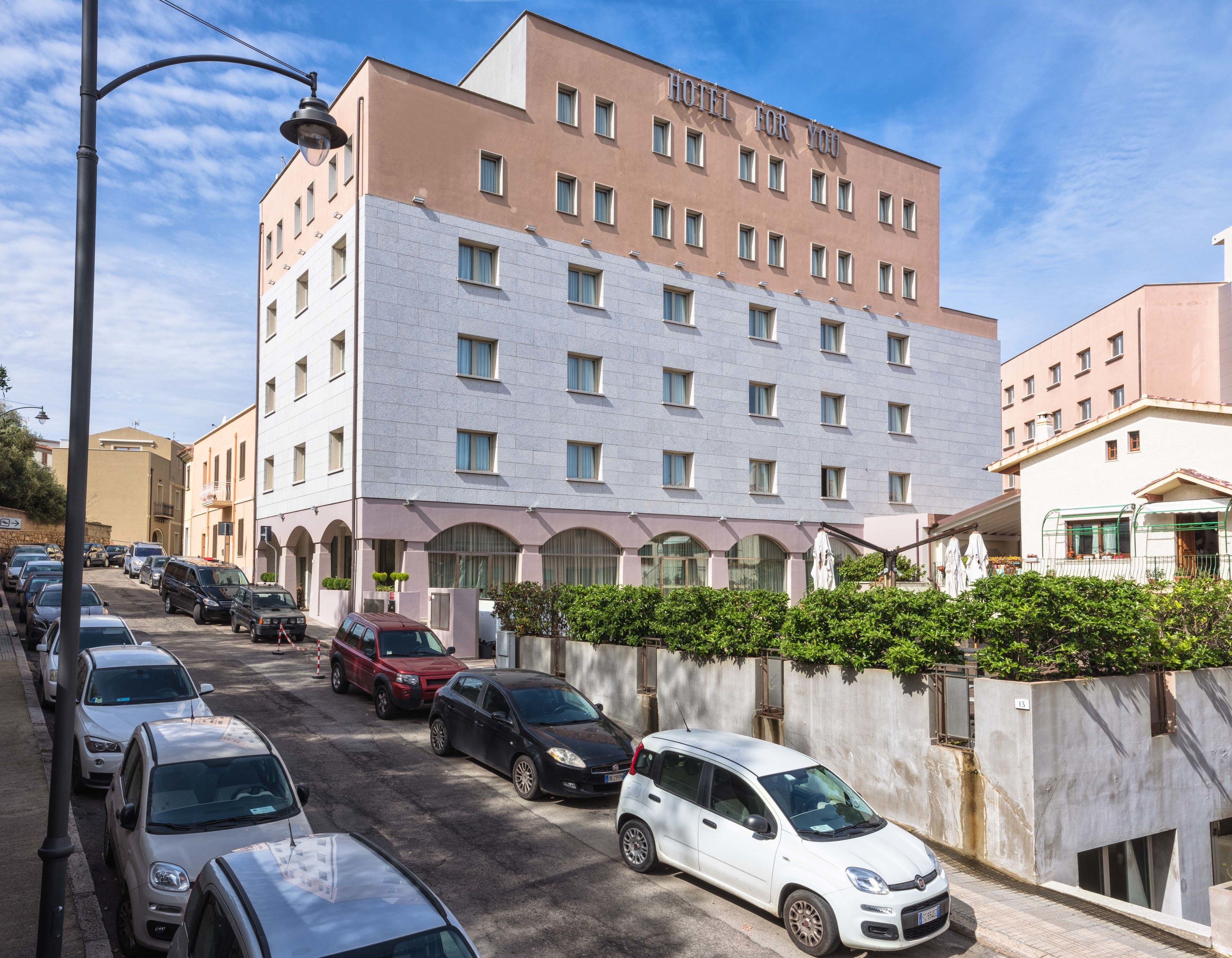 Un hotel de varios pisos con fachada moderna, un estacionamiento con varios autos y un cielo despejado.