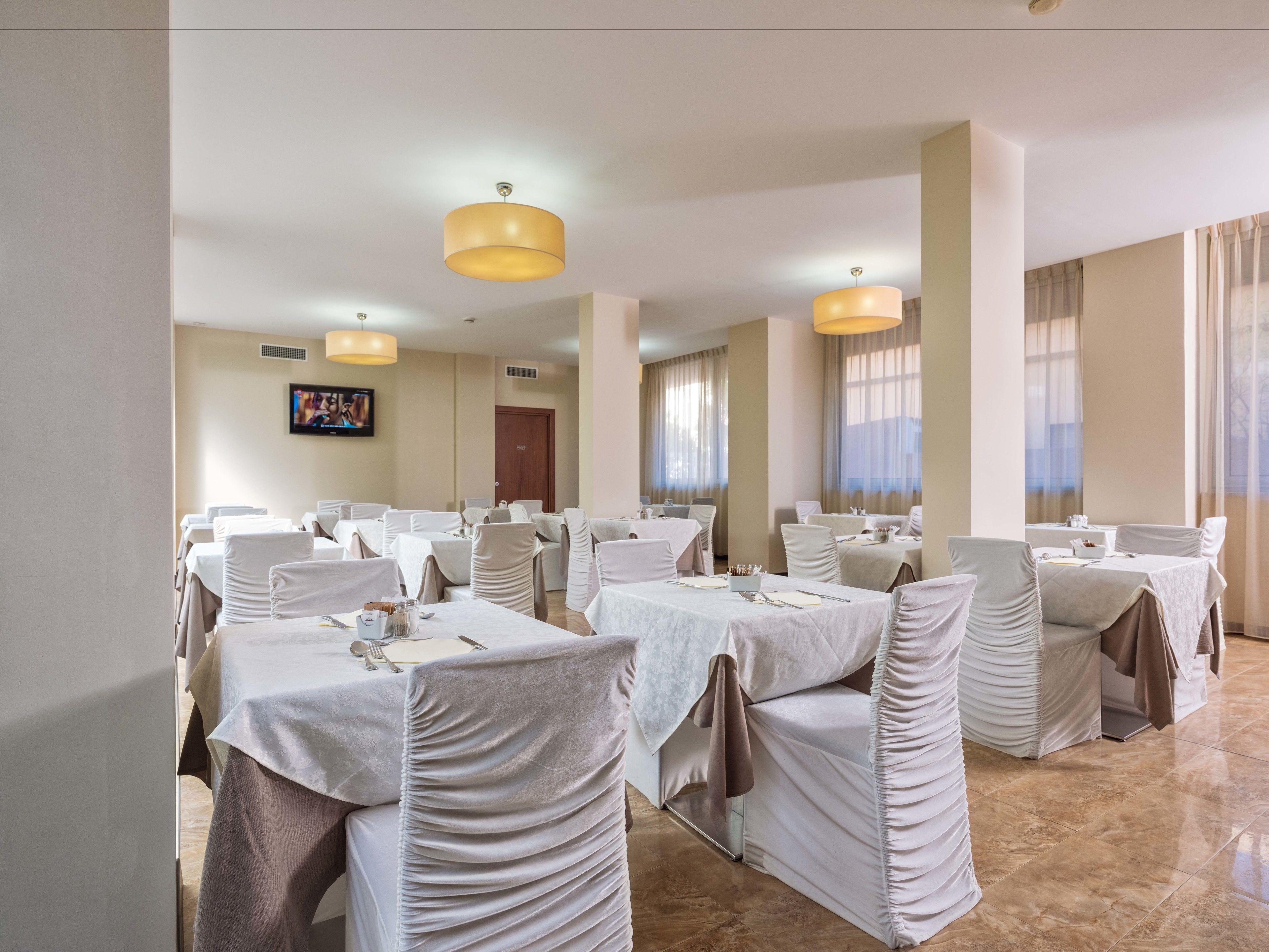 A banquet hall with multiple round tables, each covered with white tablecloths and draped with beige chair covers.