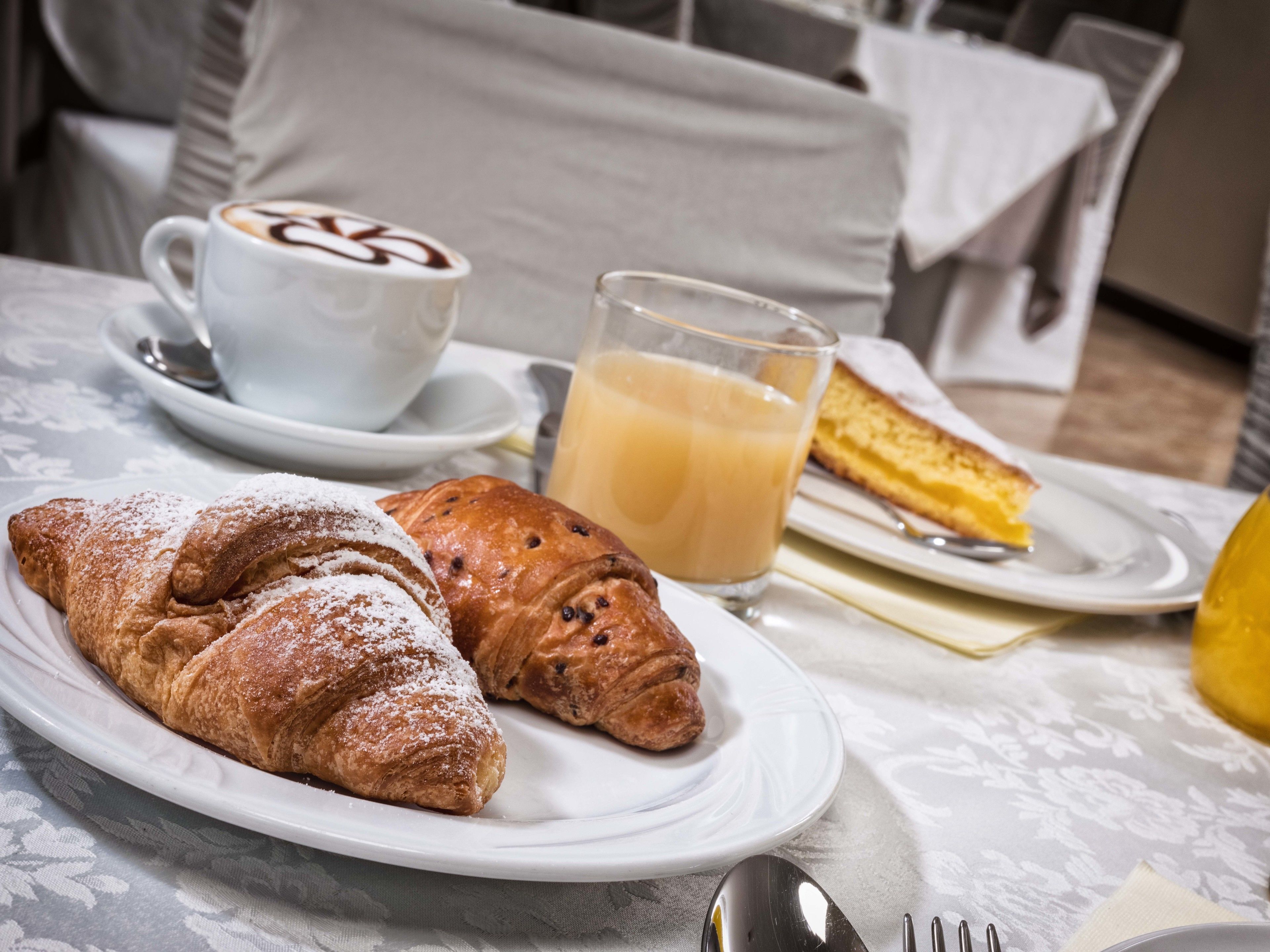 A breakfast setting with croissants, a glass of orange juice, and a slice of cake on a white tablecloth.