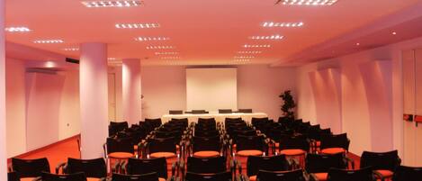Une salle de conférence avec des rangées de chaises noires et des coussins orange, un tableau blanc et un écran de projection.