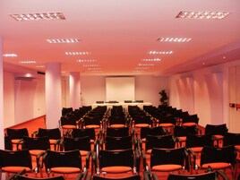 A conference room with rows of black chairs and orange cushions, a whiteboard, and a projector screen.