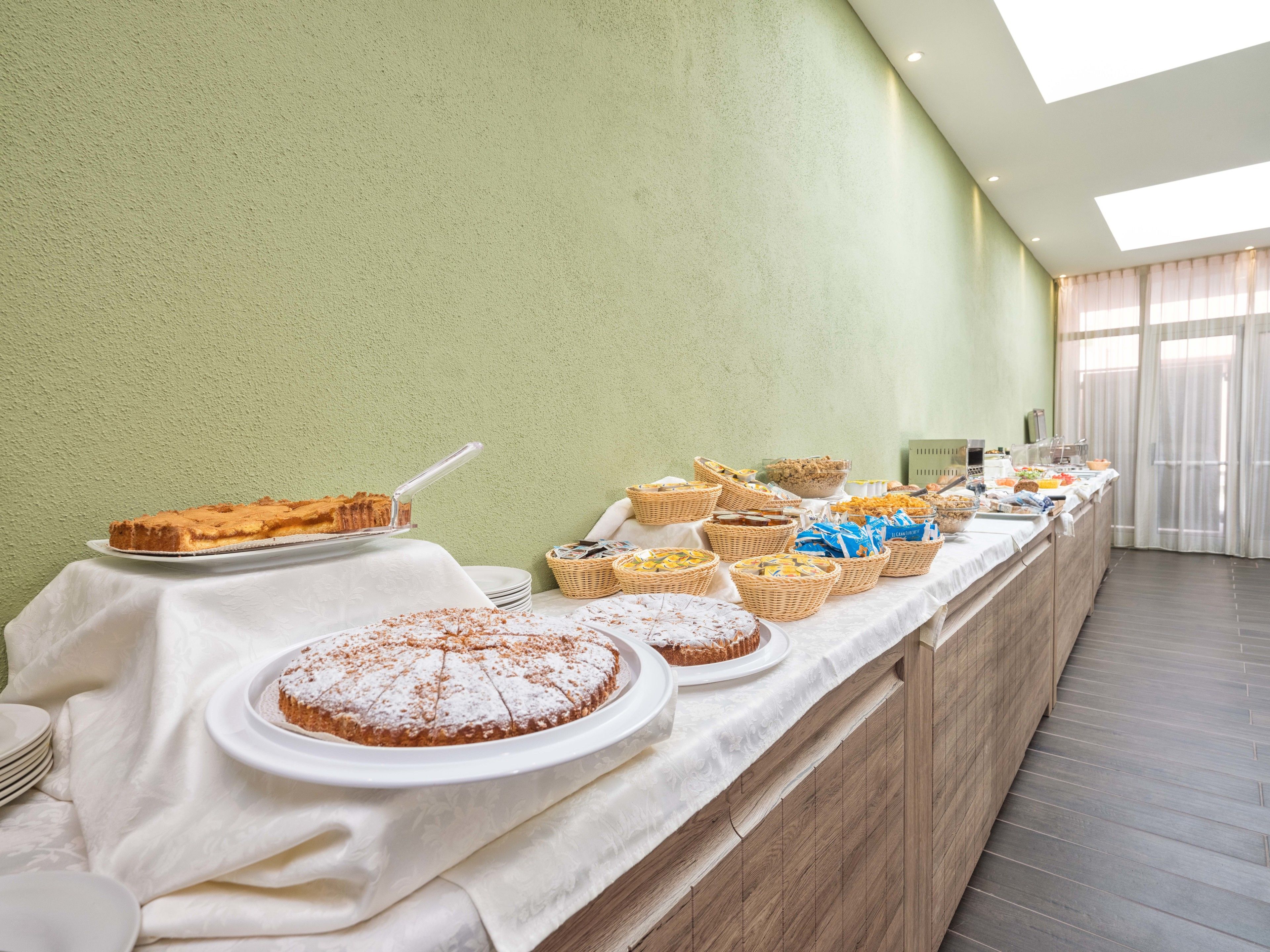 Un buffet est installé sur une table, avec une variété de desserts et de plats. Le tout est placé devant un mur vert, avec une fenêtre en arrière-plan.
