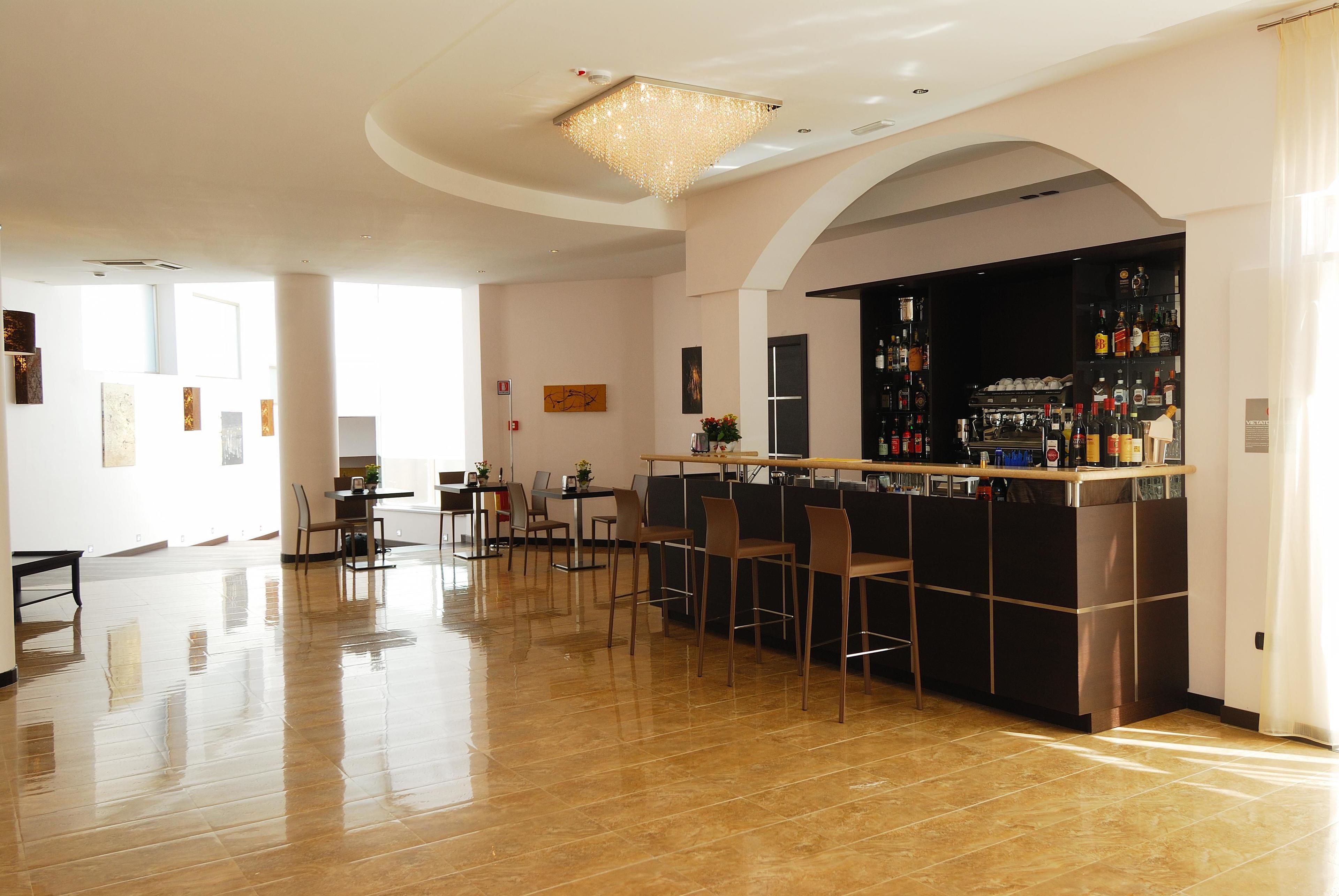 A spacious bar area with a polished floor, a bar counter, and a chandelier. There are chairs and a table in the foreground, and a door leading to another room.