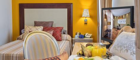 A hotel room with a bed, a bedside table with a lamp, a small desk with a phone, and a breakfast tray with fruit.