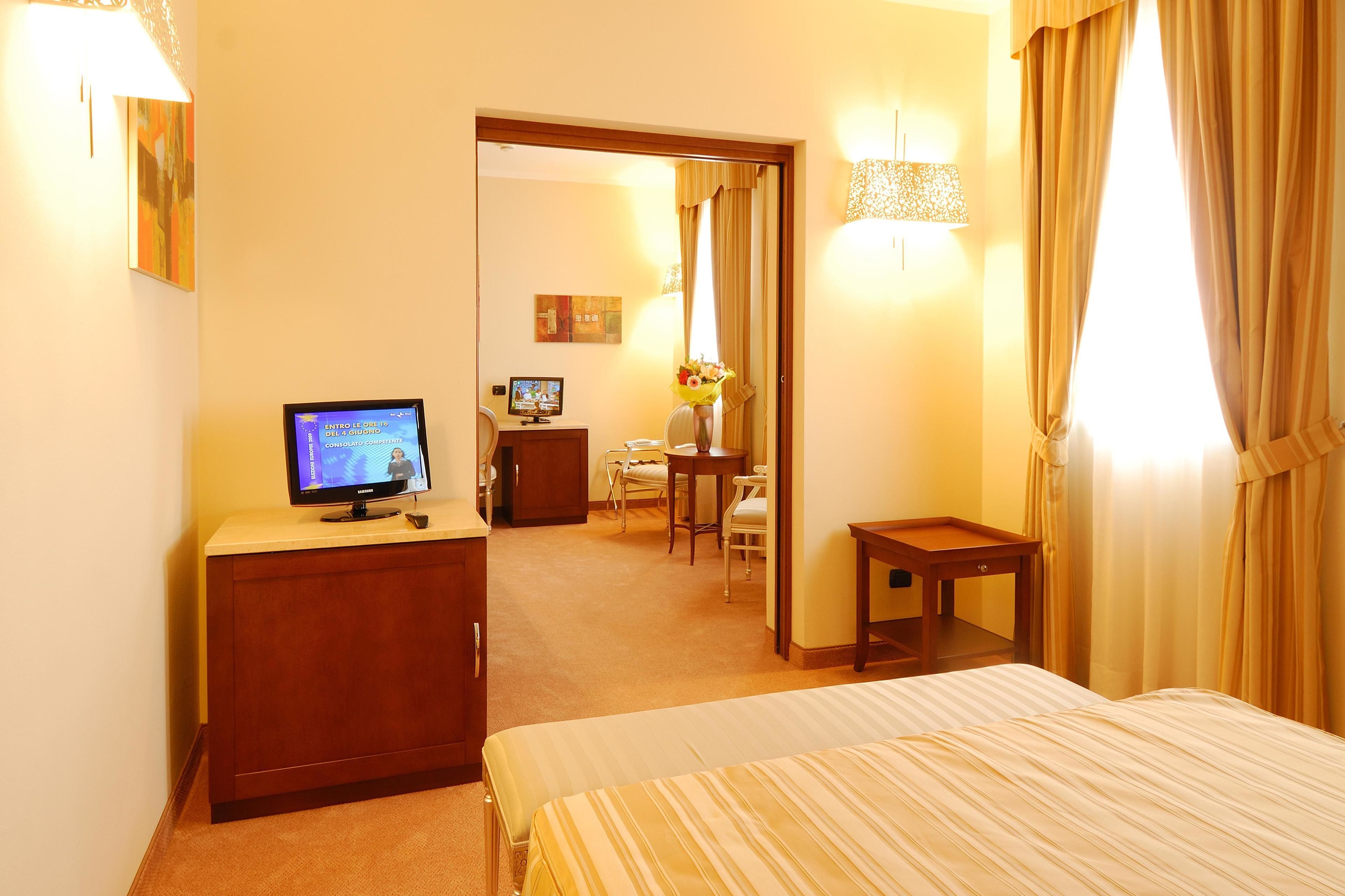 Ein Hotelzimmer mit einem Bett, einem Schreibtisch mit Computer, einem Fernseher und einem kleinen Tisch mit Blumen.