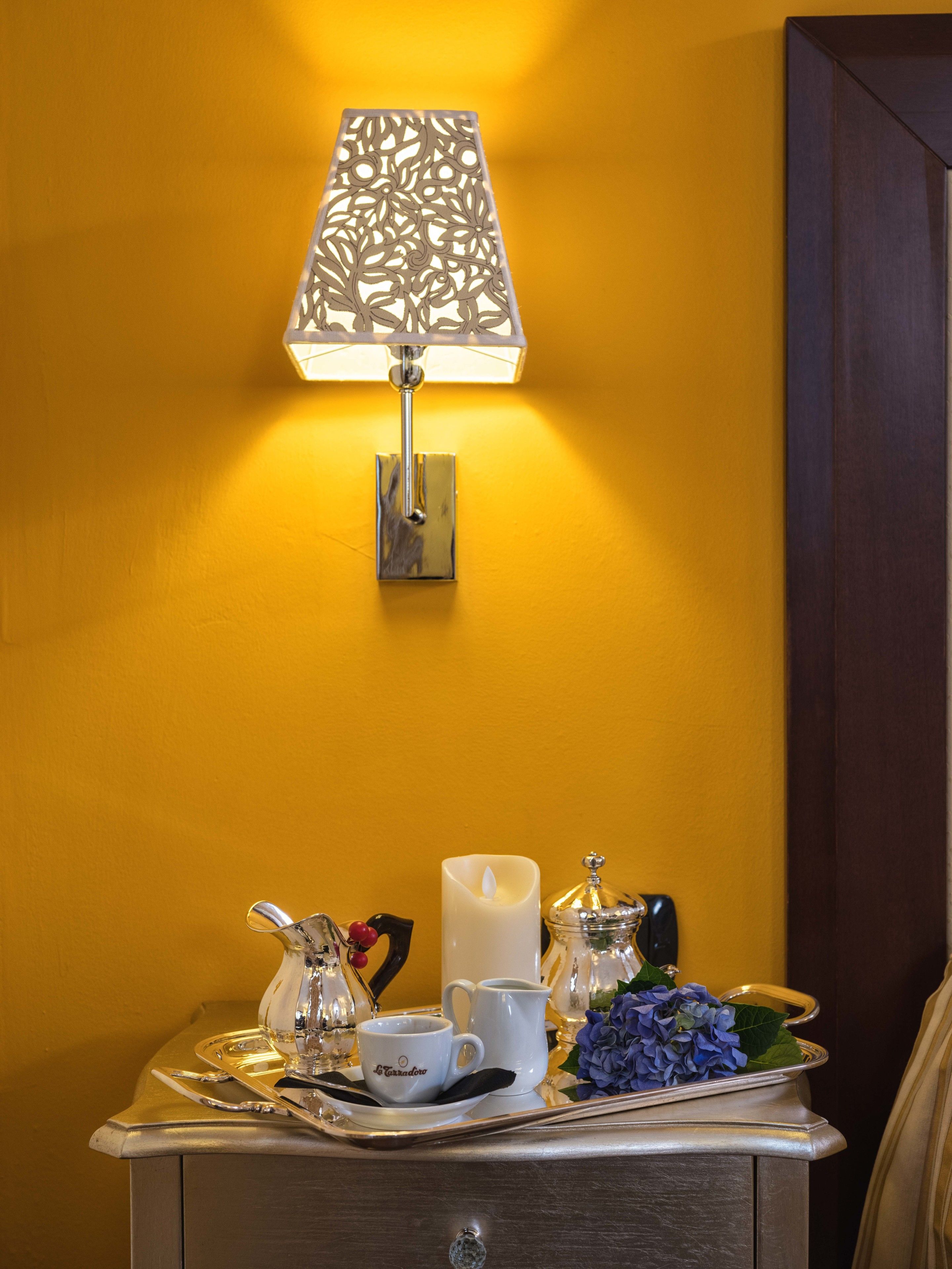 A table with a tray, a candle, a pitcher, a cup, and a vase with flowers against a yellow wall.