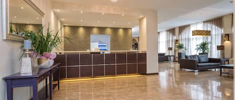 A modern hotel lobby with a reception desk, a large mirror, a potted plant, and a seating area with a sofa and armchairs.