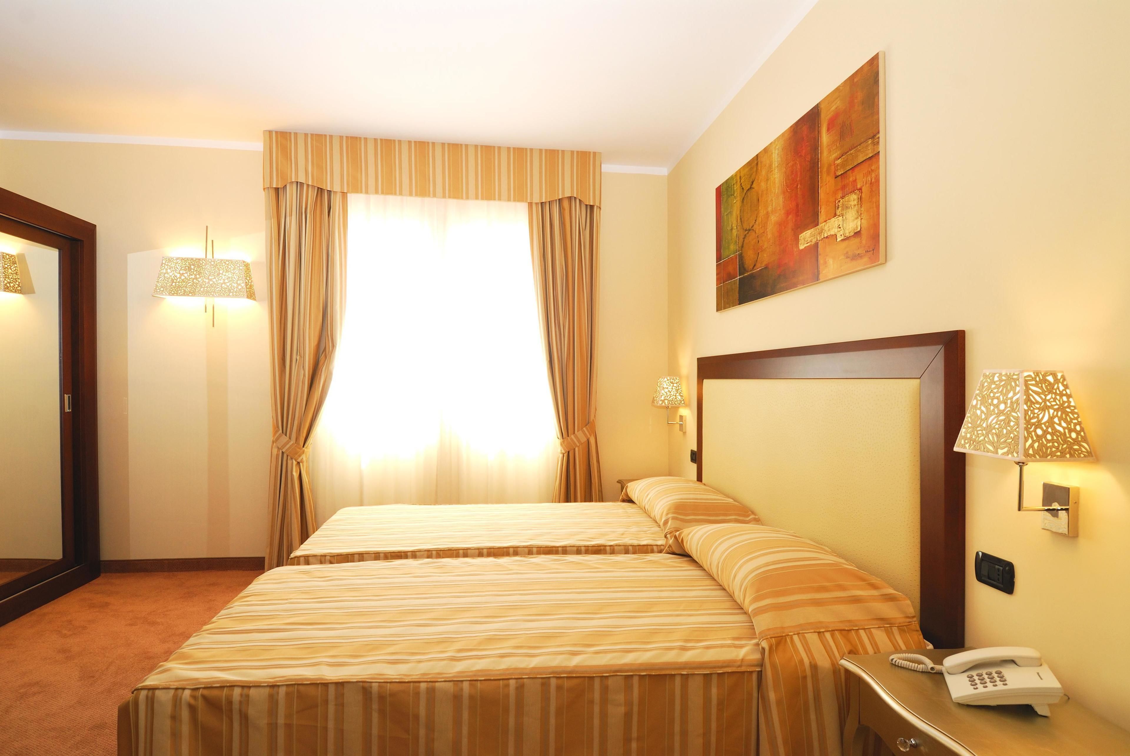 Quarto de hotel com duas camas, uma janela grande com cortinas, um quadro na parede e um telefone ao lado da cama.