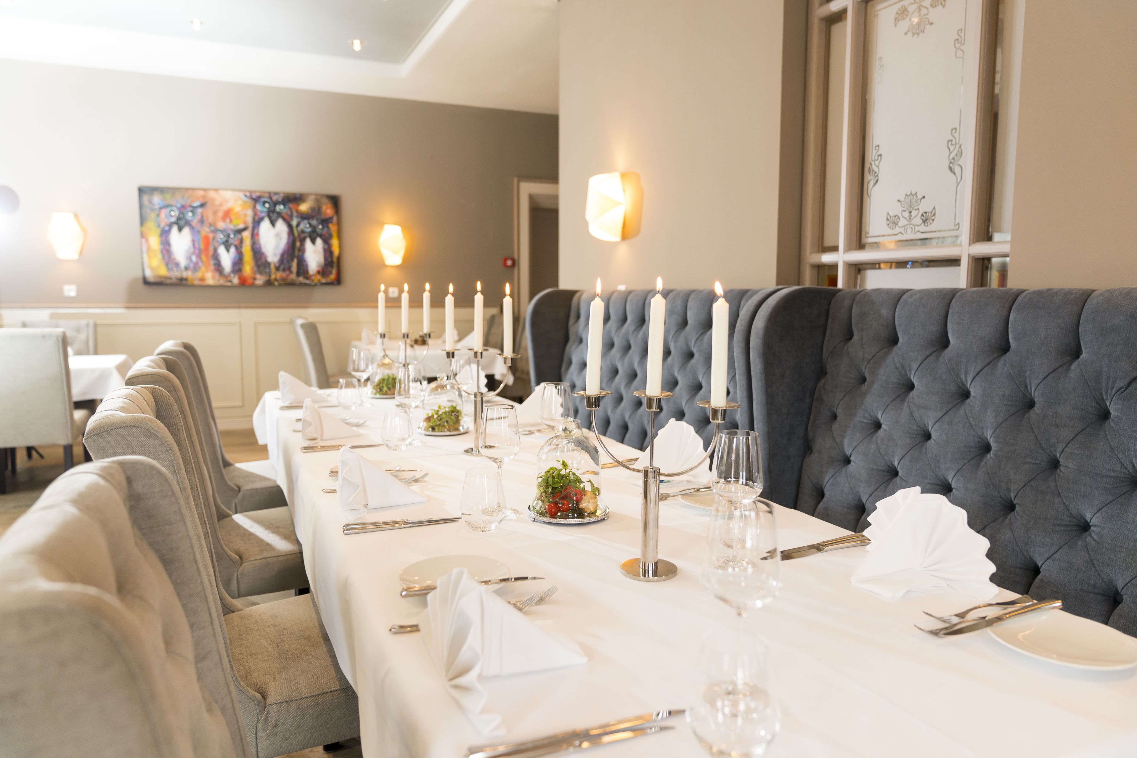 A dining area with a long table set for a meal, featuring a tufted grey upholstered bench, white tablecloths, and candle holders.