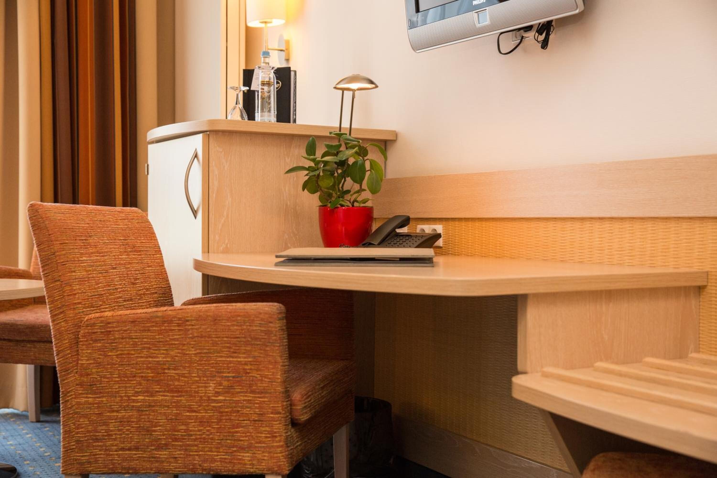 Een hotelkamer met een houten bureau, een rode plant, een lamp, een telefoon en een aan de muur bevestigde televisie.