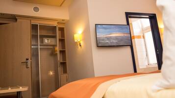 A hotel room with a bed, a wardrobe, a TV mounted on the wall, and a window with a view of the beach.