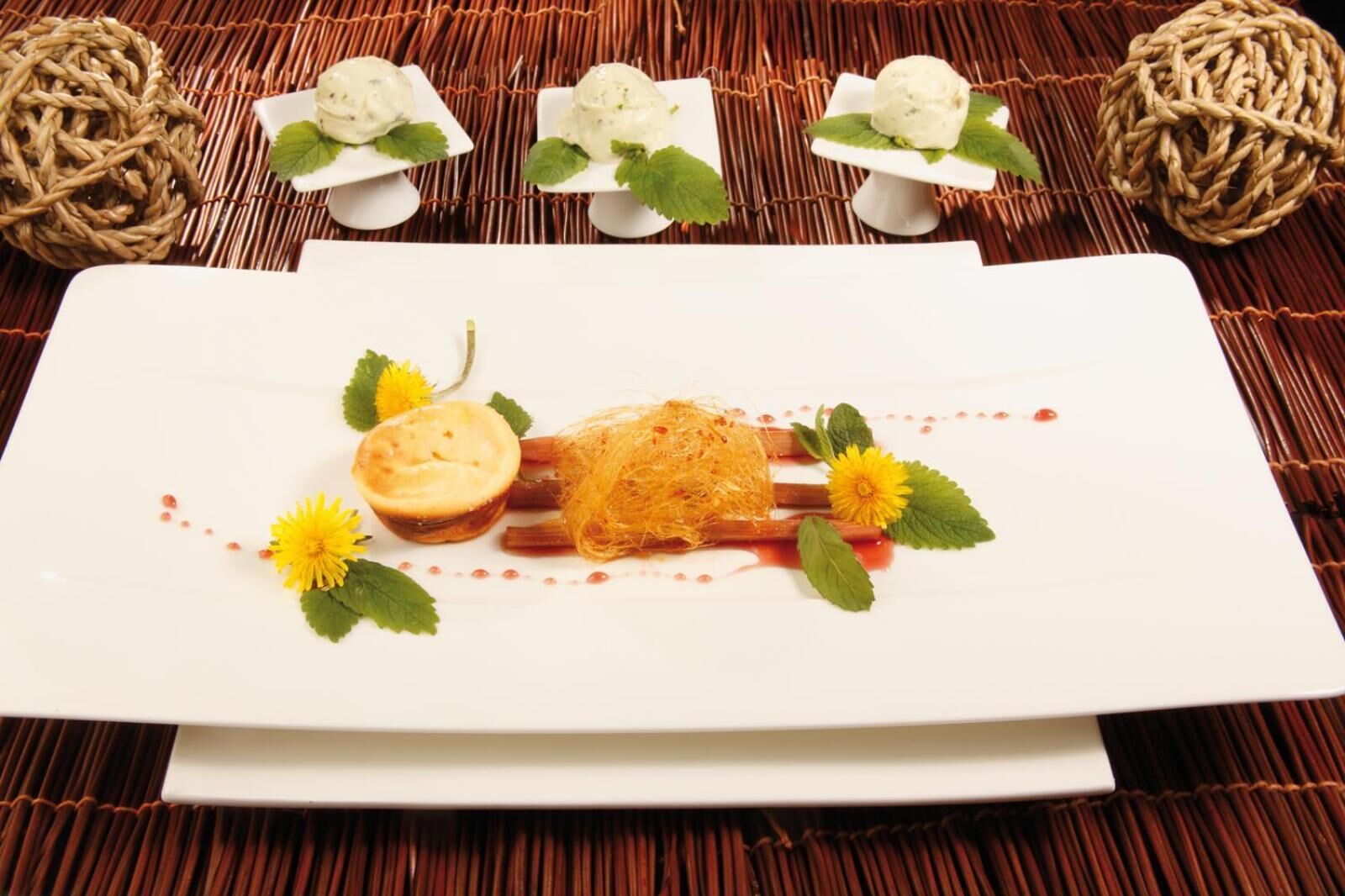 Un plato de postre con un pastel, hojas de menta y una flor amarilla sobre una fuente blanca.