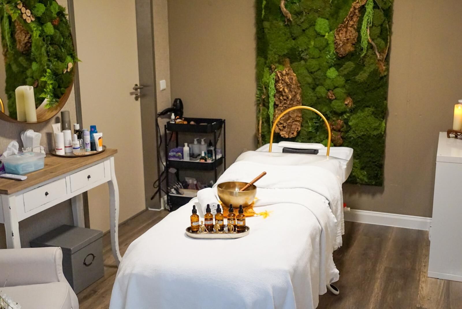 Una sala de spa con una camilla de masaje, un mostrador de madera con varias botellas, un estante con más botellas y una pared decorada con follaje verde.