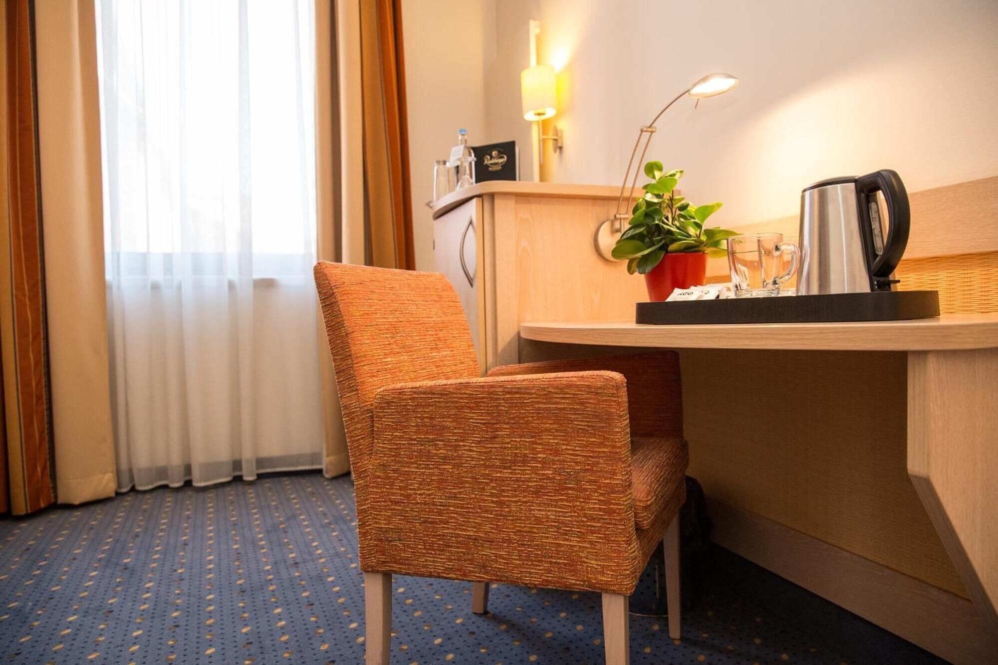 A hotel room with a woven chair, a desk with a coffee maker, a potted plant, and a lamp.