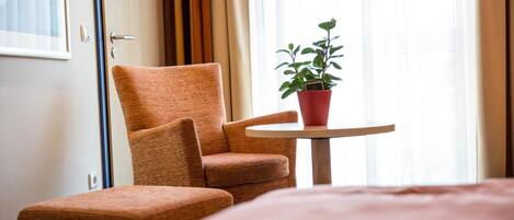 A hotel room with a bed, a chair, a small table, and a potted plant.
