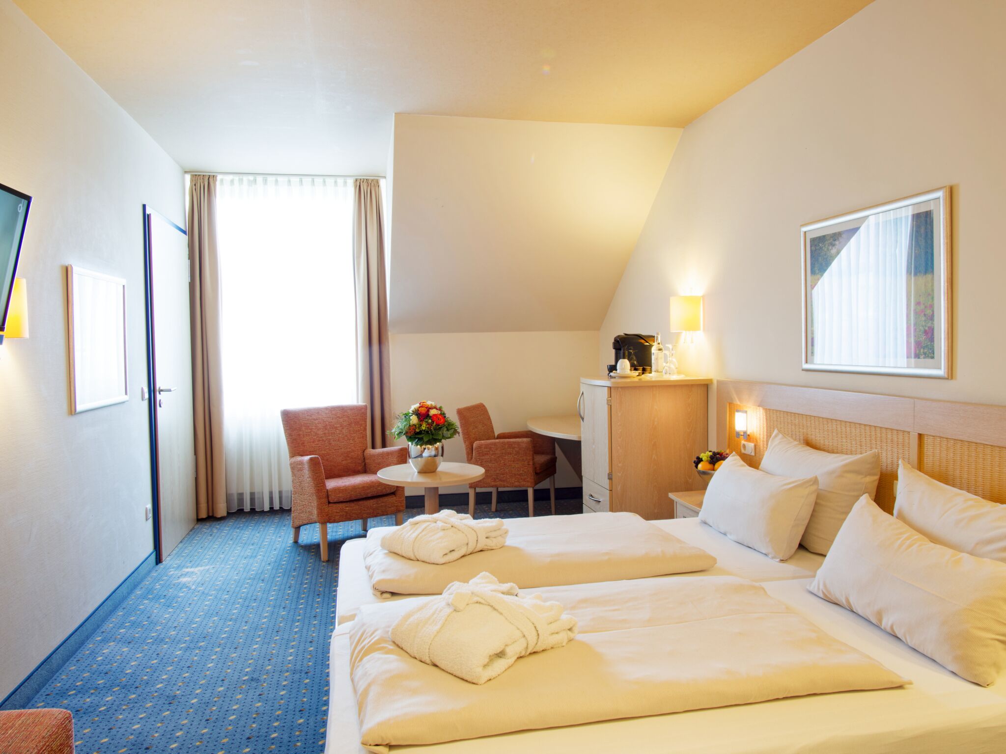 A hotel room with two beds, a desk, a chair, and a small table with a vase of flowers.