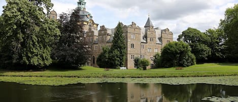A large, historic building with multiple towers and a pond in the foreground.