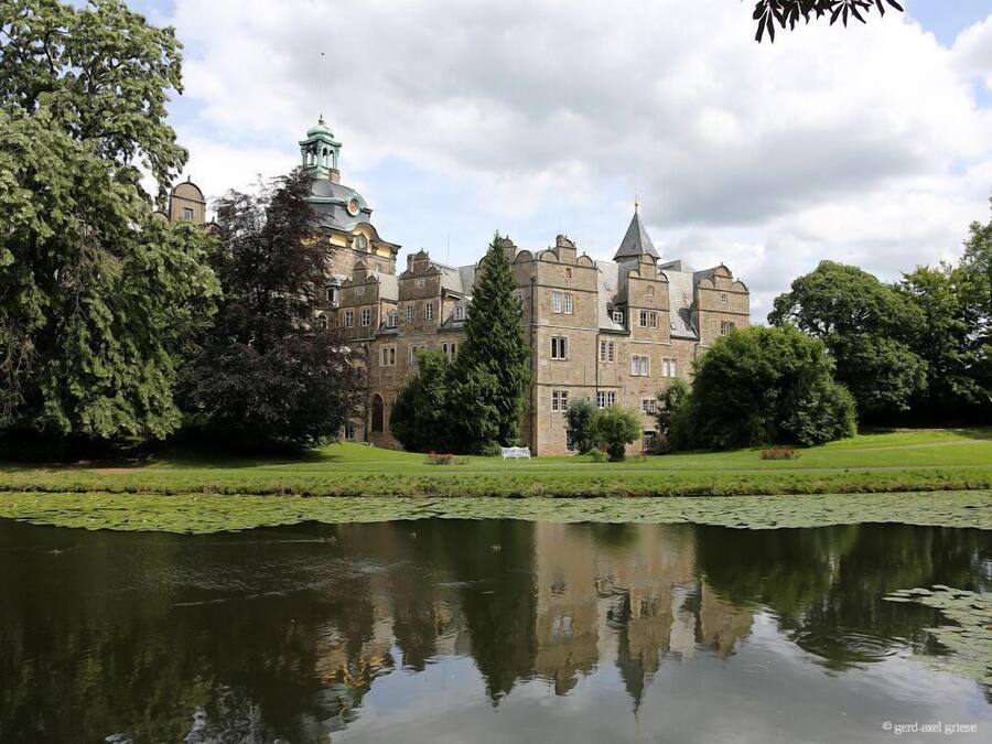 A large, historic building with multiple towers and a pond in the foreground.