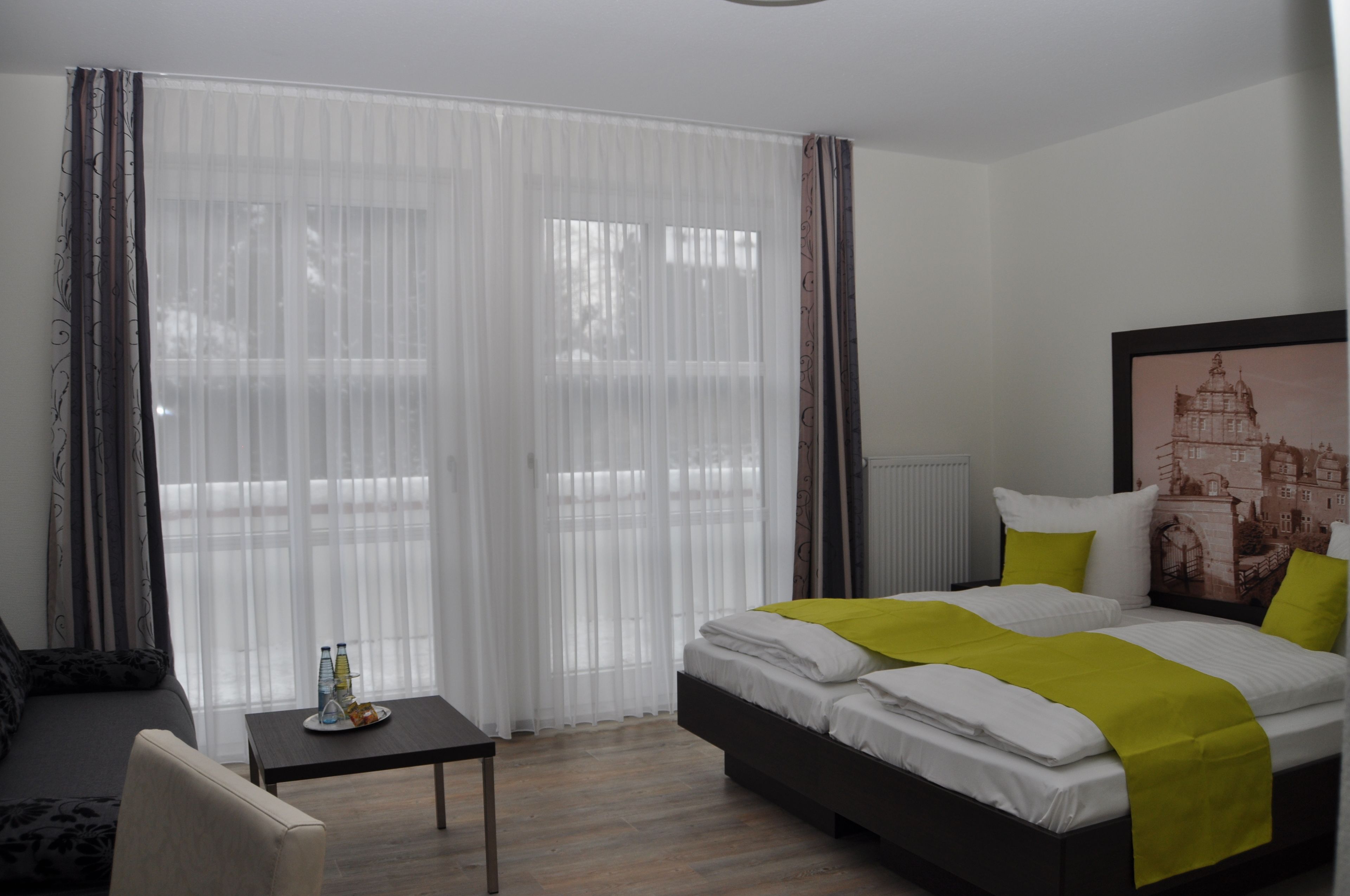 Ein Hotelzimmer mit einem Bett, einem kleinen Tisch mit Flasche und Gläsern, einem Sessel und einem großen Fenster mit durchsichtigen Vorhängen.