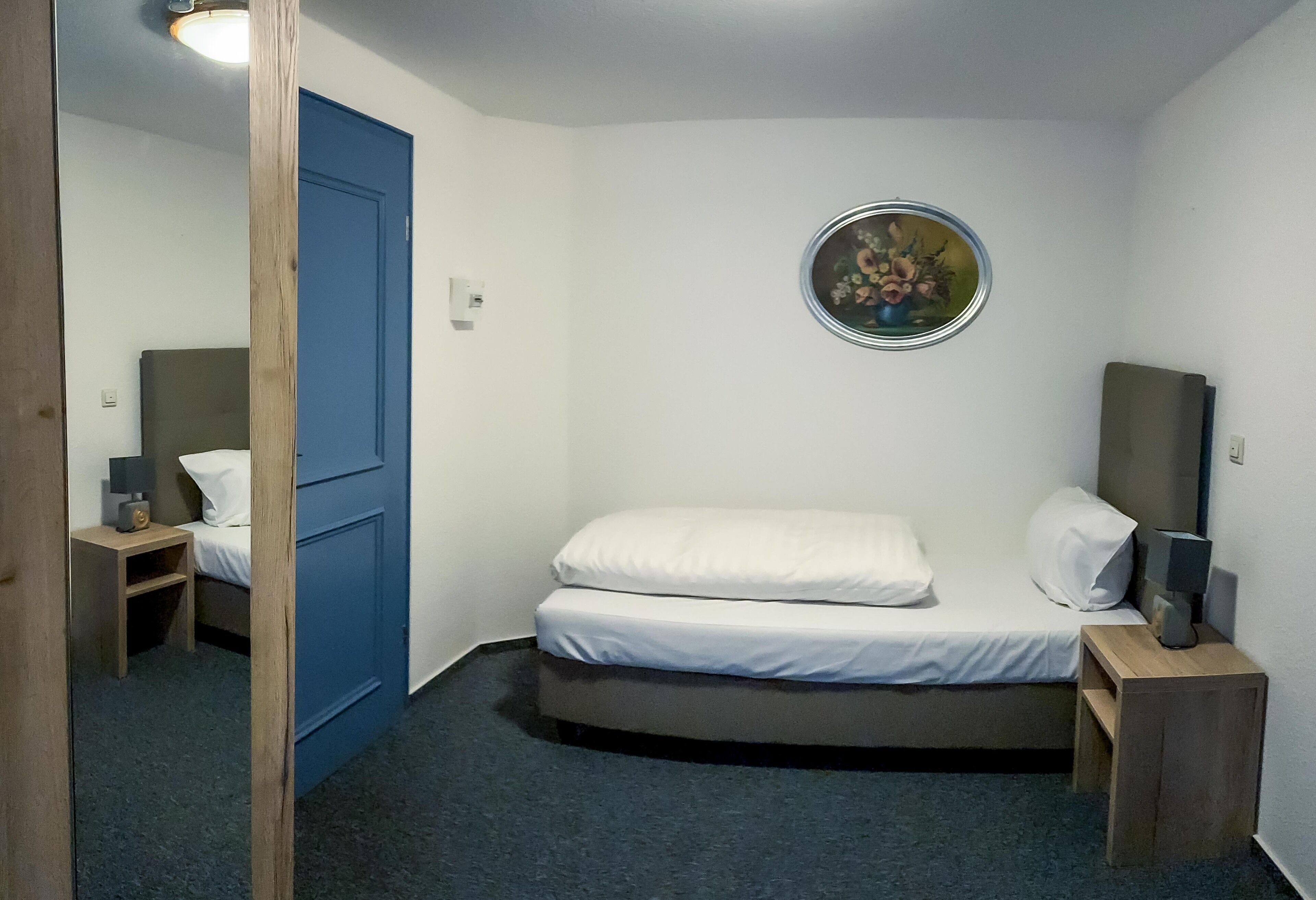 A small, compact room with a single bed, a bedside table, a lamp, and a wall-mounted picture.