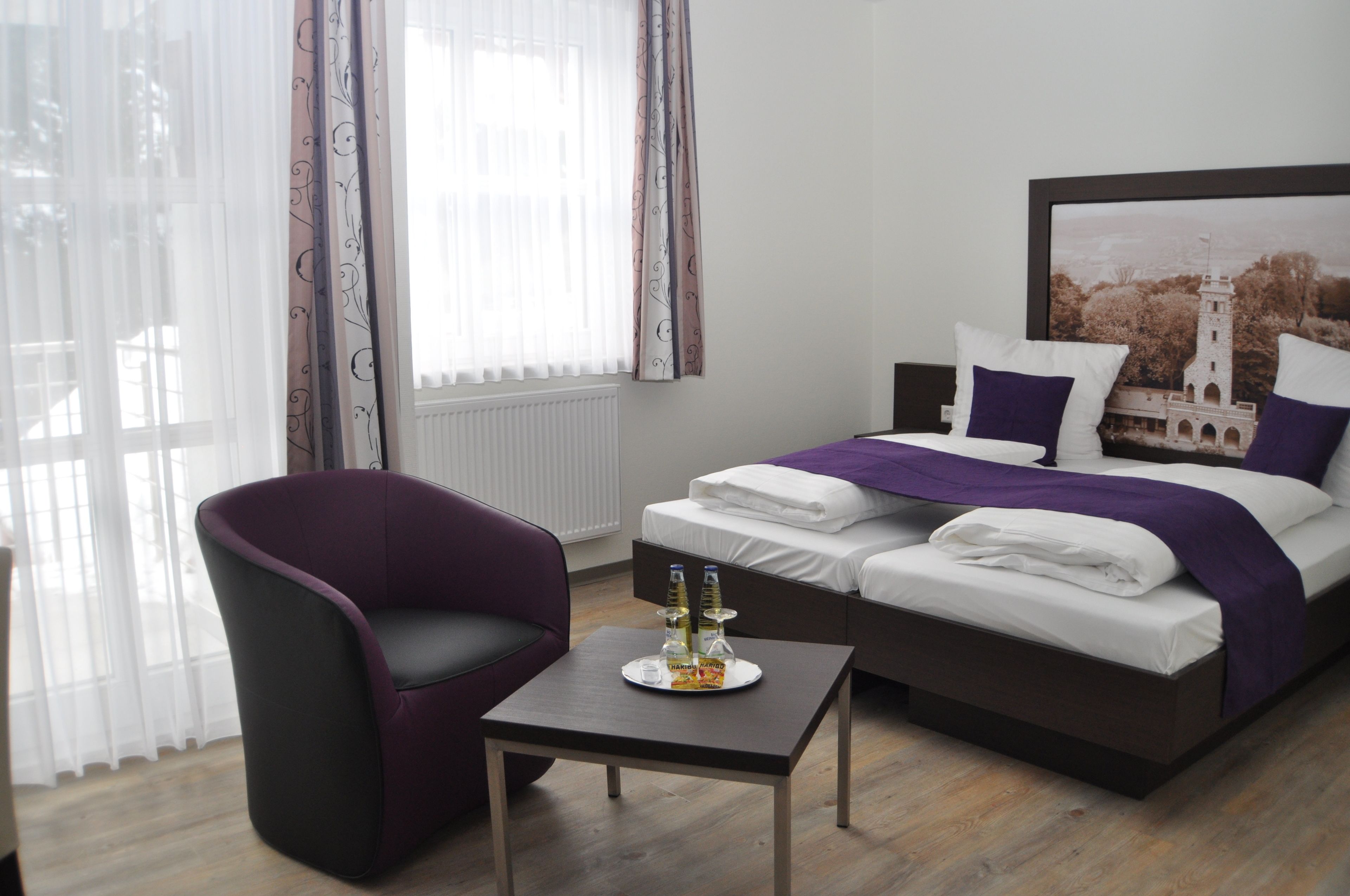 Ein modernes Hotelzimmer mit Bett, Sessel, kleinem Tisch und einem Vorhangfenster.
