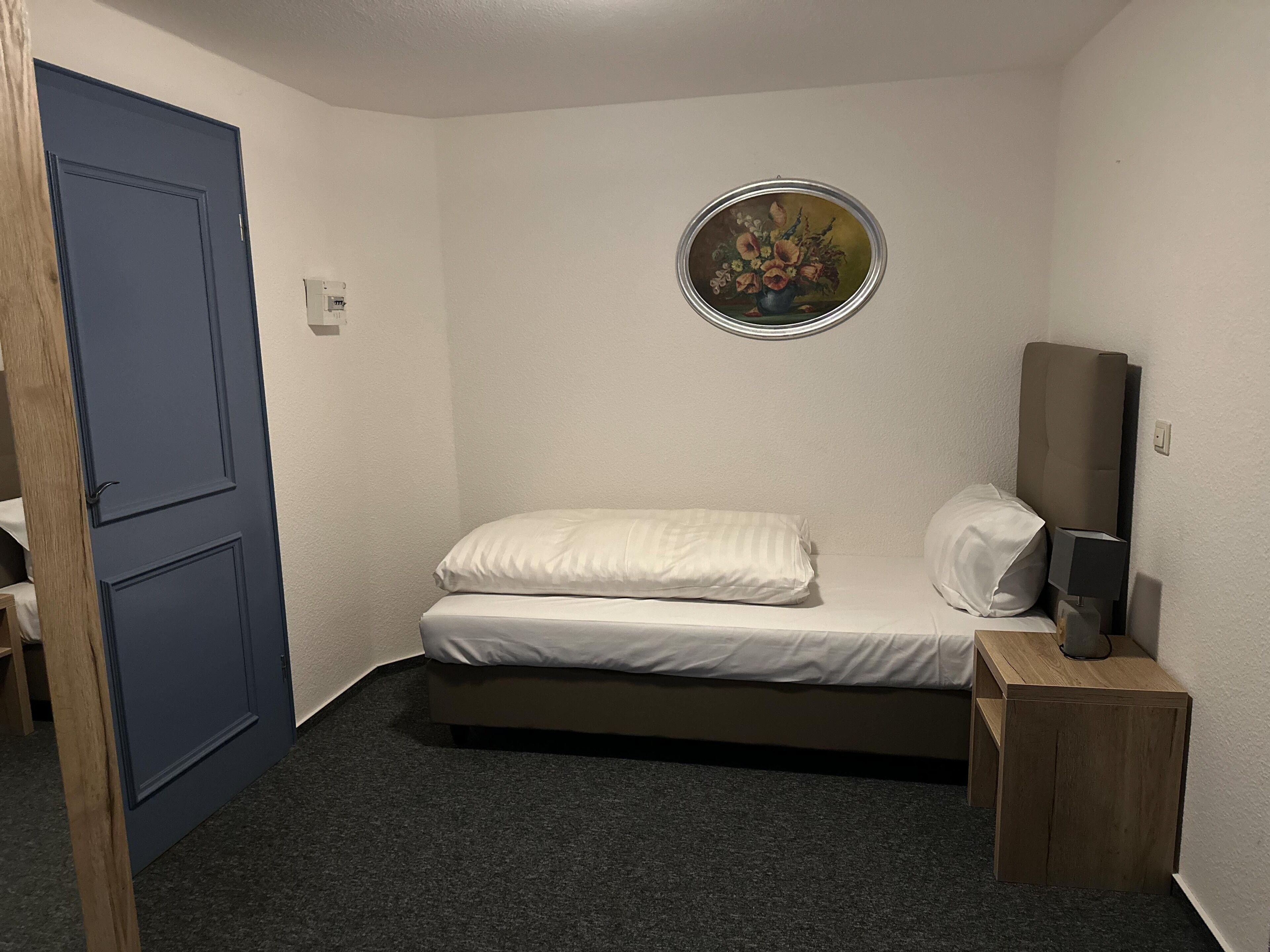 Une petite chambre à coucher, bien rangée, avec un lit simple, une table de chevet, un miroir et un tableau accroché au mur.