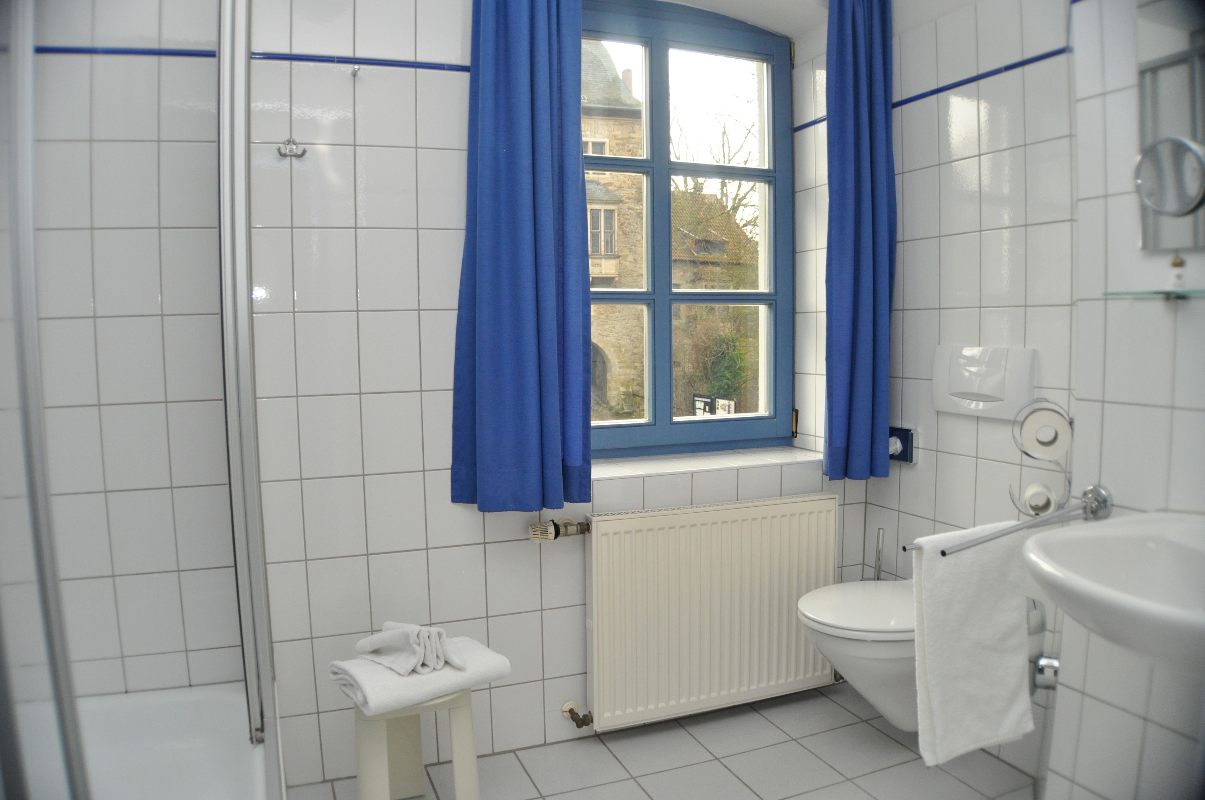 Ein Badezimmer mit einem weißen Waschbecken, einer Toilette und einer Badewanne. Ein Fenster mit blauen Vorhängen bietet einen Blick auf ein Gebäude.