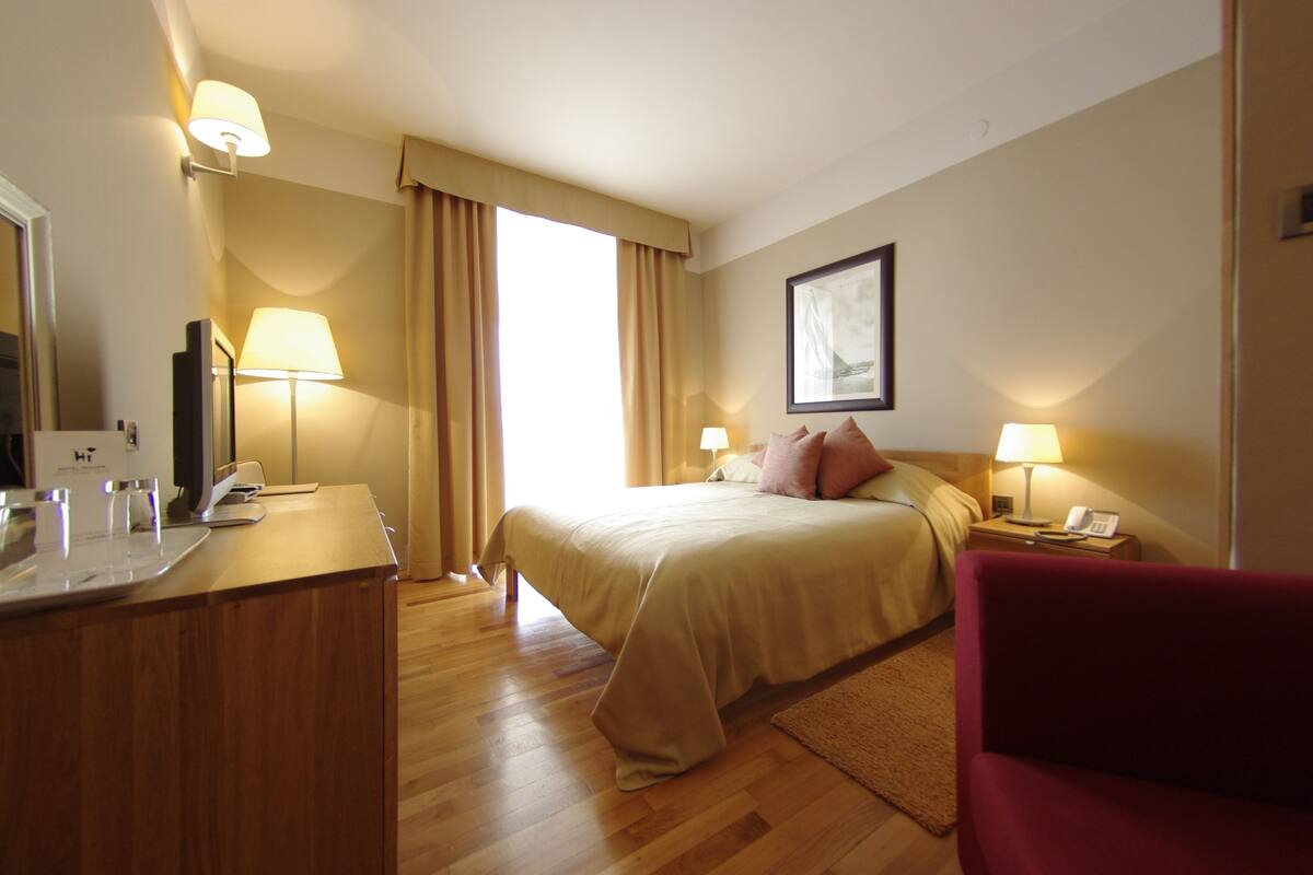 Habitación de hotel con cama, lámparas de noche, escritorio con lámpara, silla y un ventanal con cortinas.