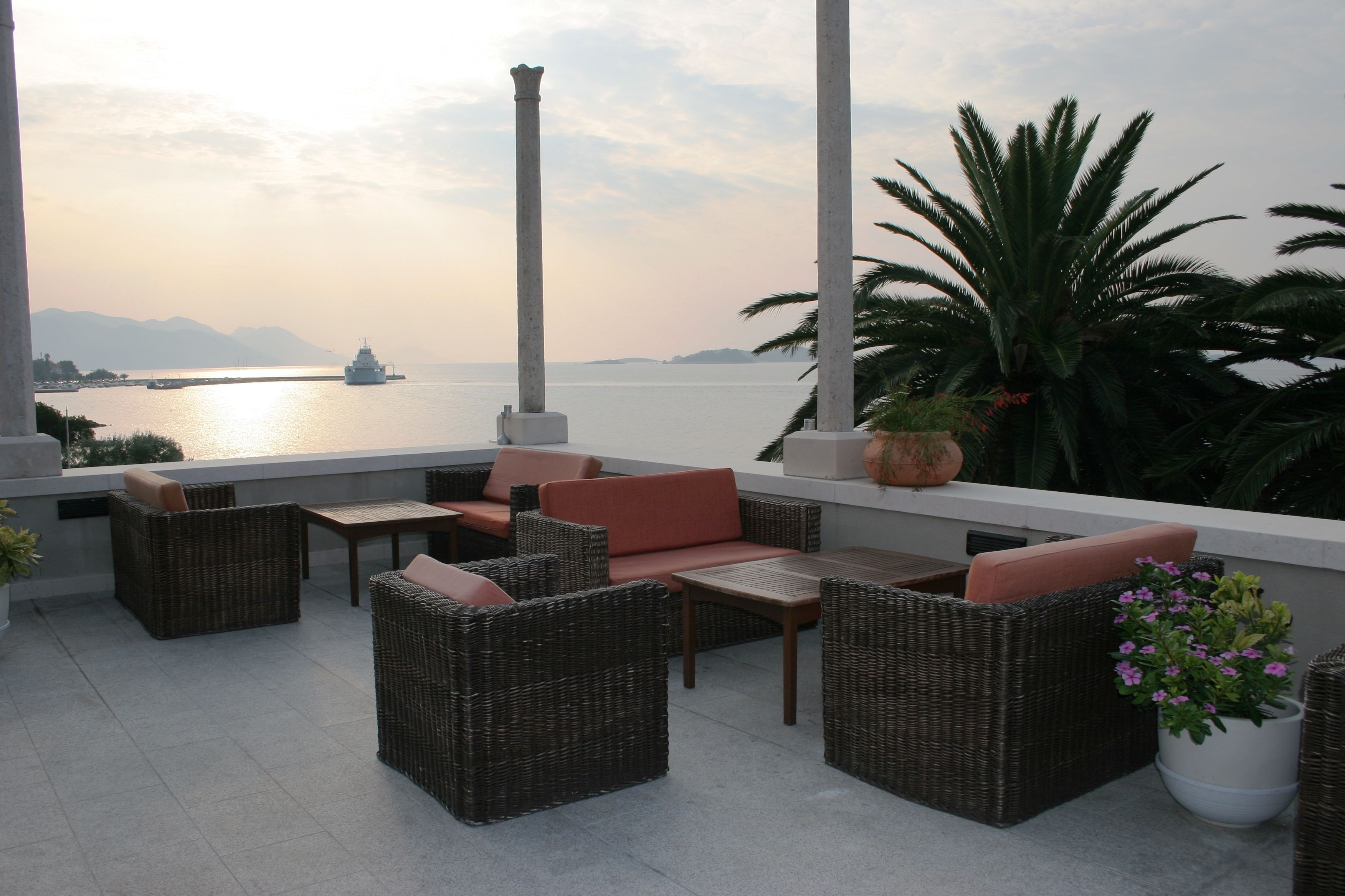 A terrace with wicker furniture, a view of the sea, and a ship in the distance.