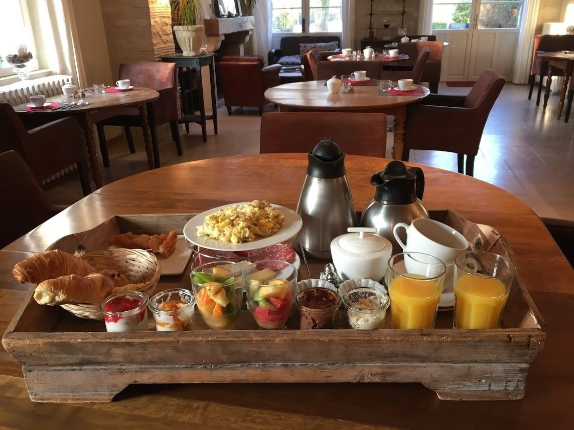 Un vassoio per la colazione con croissant, frutta, yogurt e caffè su un tavolo di legno in sala da pranzo, con sedie e tavoli.