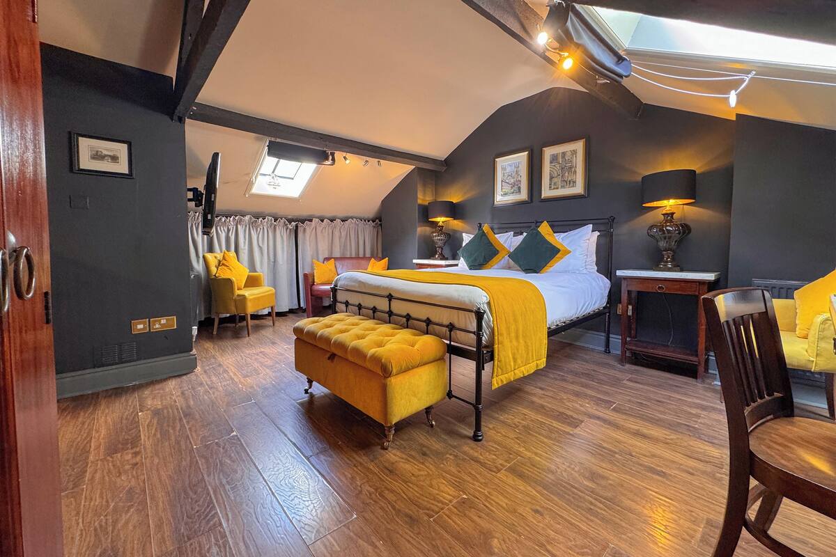 A bedroom with a bed, a yellow armchair, a wooden bench, a desk, and a window with curtains.