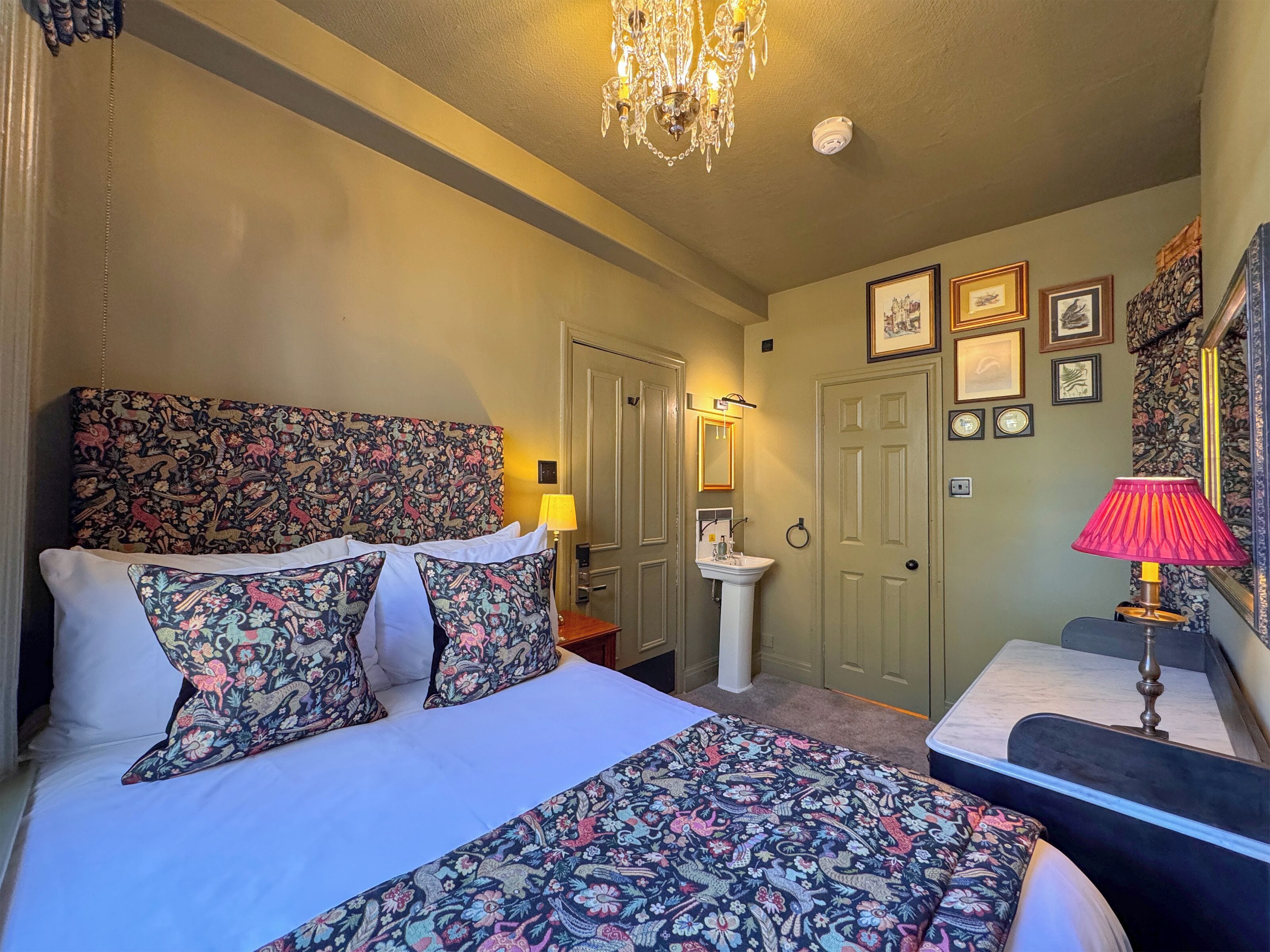 A bedroom with a floral-patterned headboard, a bedside table with a lamp, a white sink, and a door with a mirror.