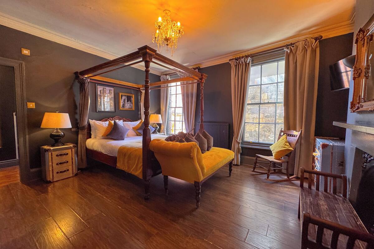 A four-poster bed, a wooden floor, a chair, a lamp, and a window with curtains.
