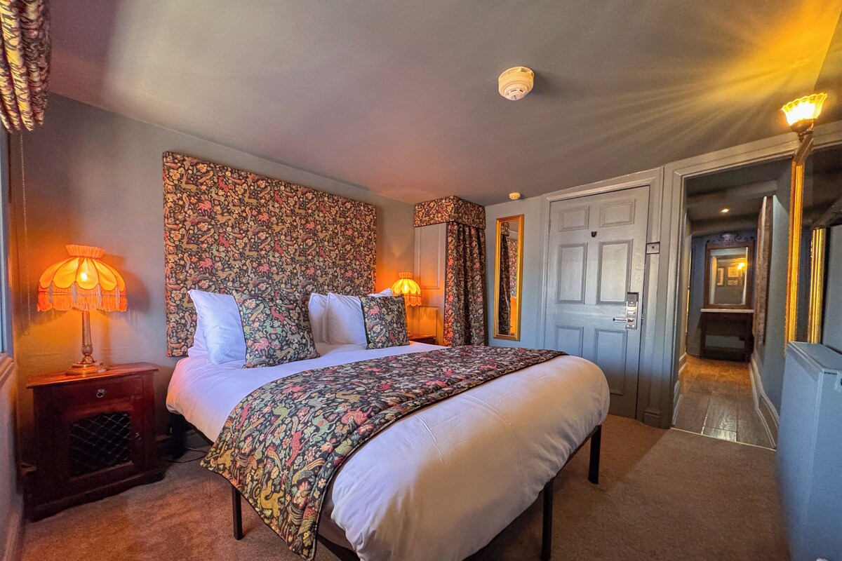 Una habitació d'hotel amb un llit gran, dues làmpades de nit, un capçal de llit floral, un mirall i una porta que dona a una altra habitació.