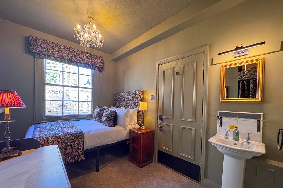 A bedroom with a bed, bedside table, lamp, window with patterned curtains, chandelier, door, and a bathroom with a sink and mirror.