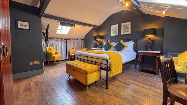 A bedroom with a bed, a yellow armchair, a wooden bench, a desk, and a window with curtains. - The Fat Badger (York)