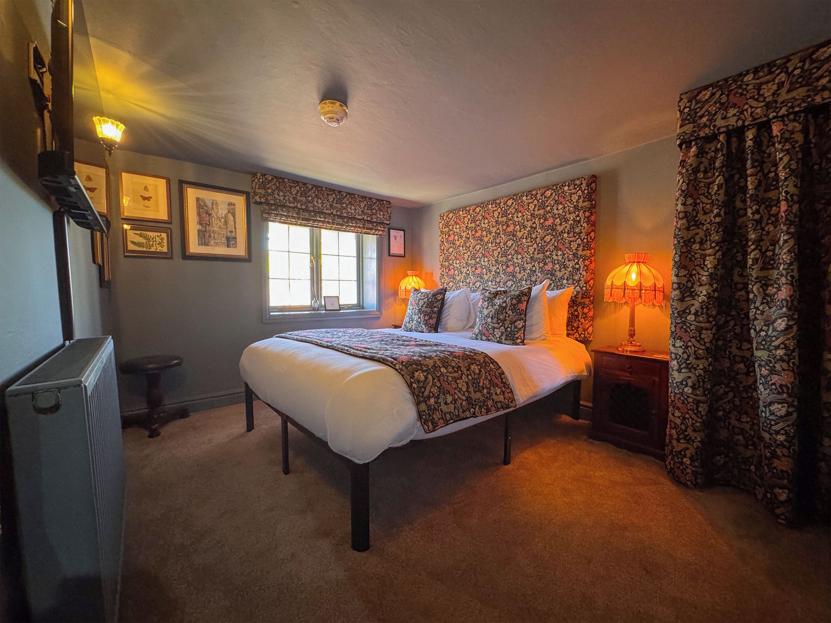 A bedroom with a large bed, two bedside lamps, a radiator, a window with a view, and patterned curtains.