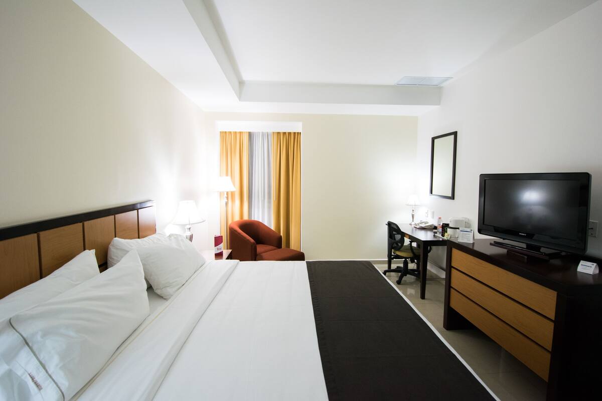 Une chambre d’hôtel avec un lit, un bureau, une chaise, une télévision et une fenêtre avec des rideaux.