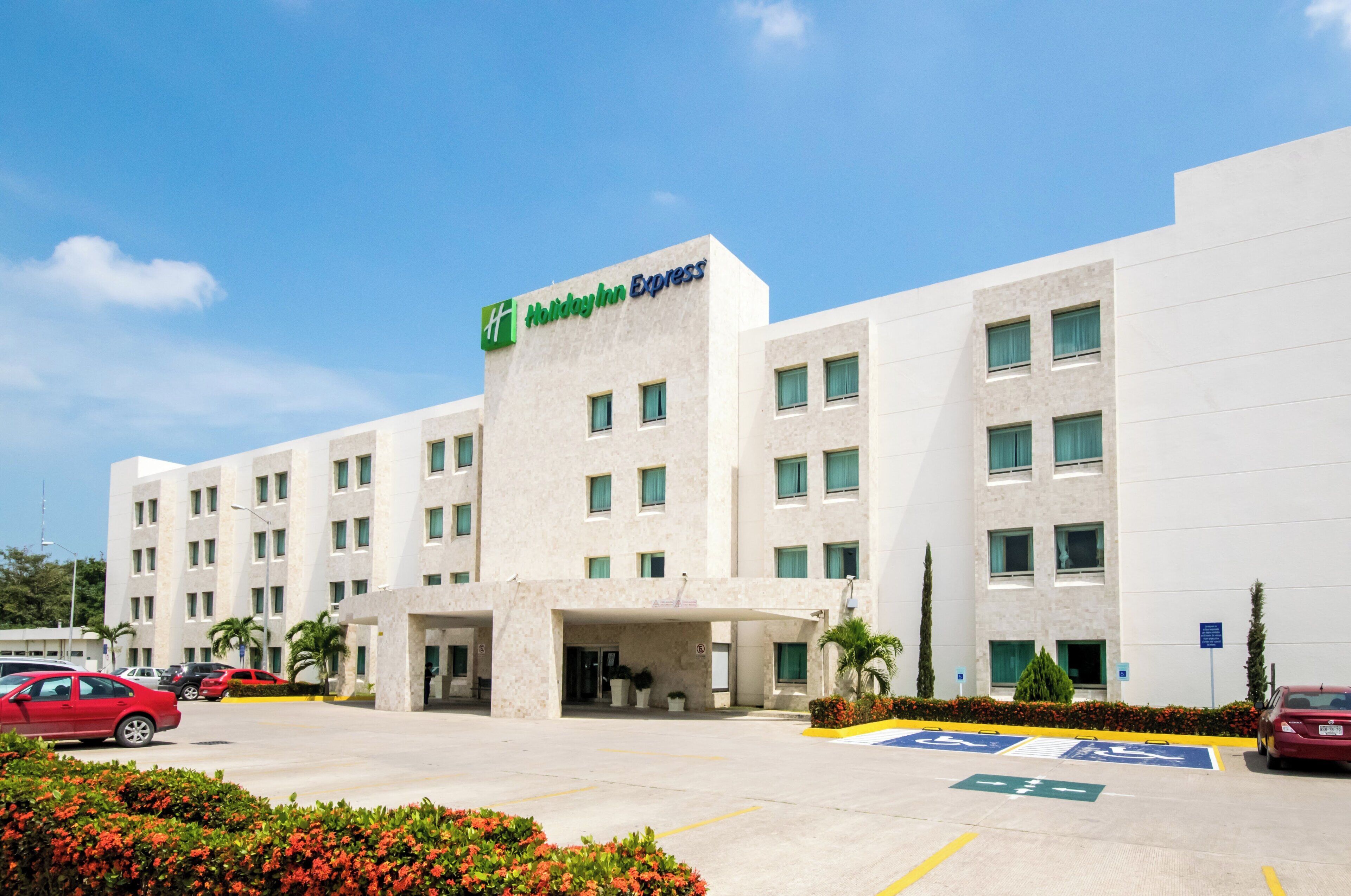Un hotel Holiday Inn Express con fachada moderna, varios ventanales y un estacionamiento con espacios reservados para discapacitados.