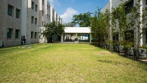 A courtyard with a building featuring a covered seating area, surrounded by trees and a grassy lawn.