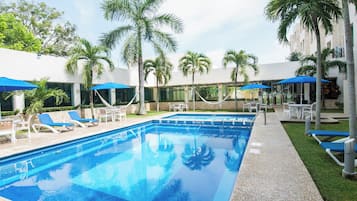 A swimming pool with lounge chairs and umbrellas, surrounded by palm trees and a building.