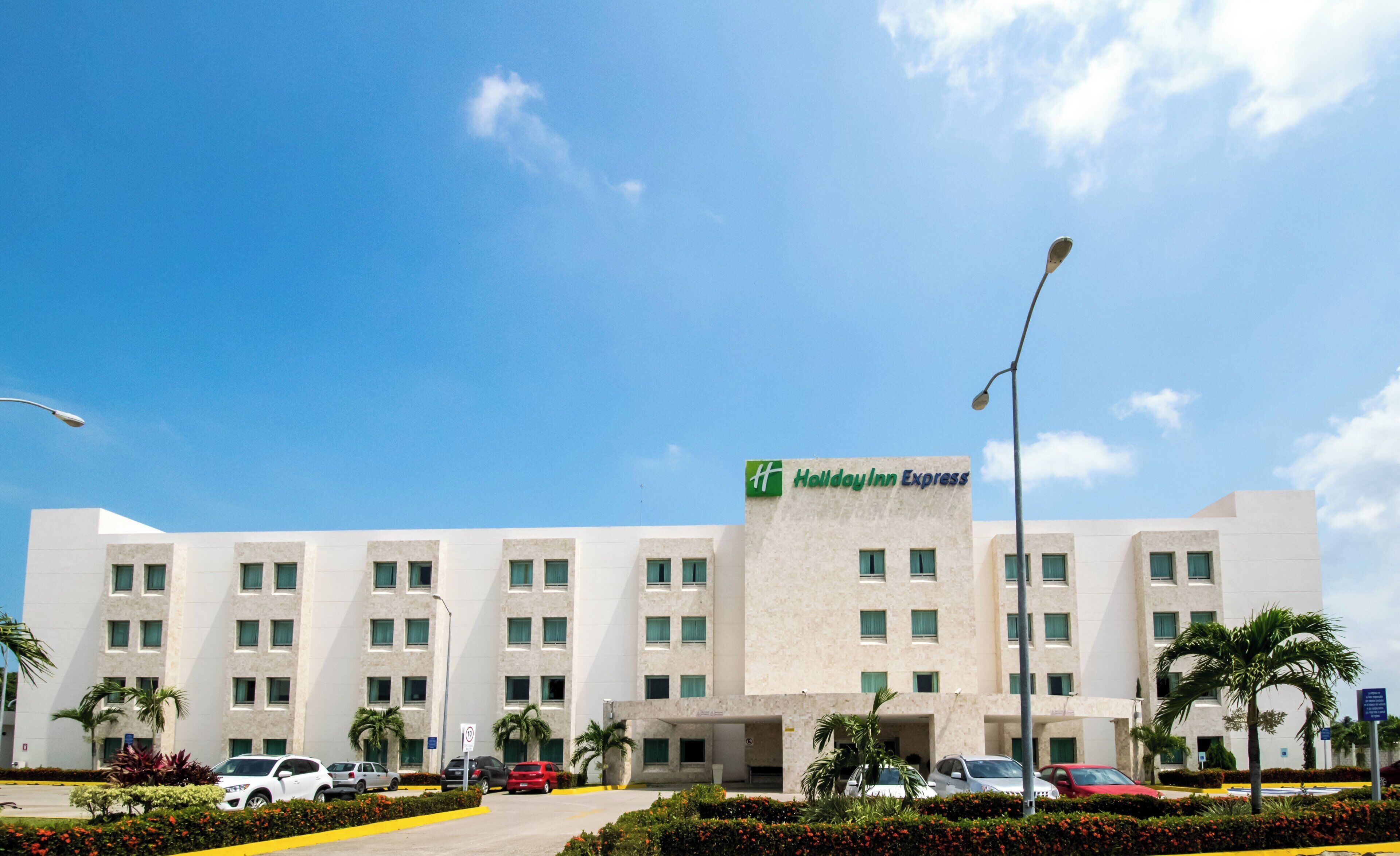 Un hotel de varios pisos con fachada plana, entrada visible y estacionamiento con varios automóviles.